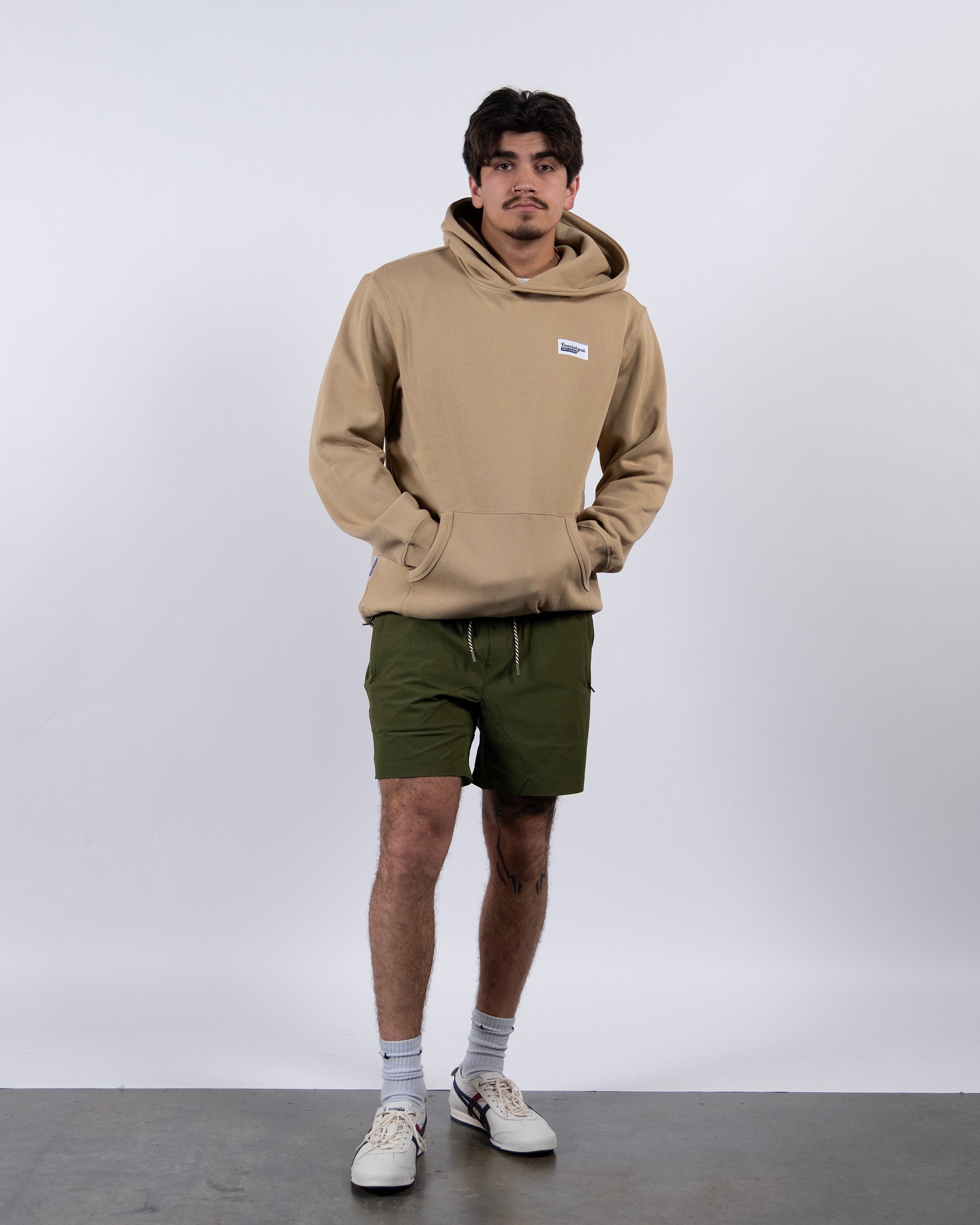 A man with dark hair and a mustache stands against a white background wearing beardedgoat’s best-selling The VOID™ Hoodie made from heavyweight cotton fleece, paired with olive green shorts, white socks, and white sneakers. His hands are in the hoodie pocket.