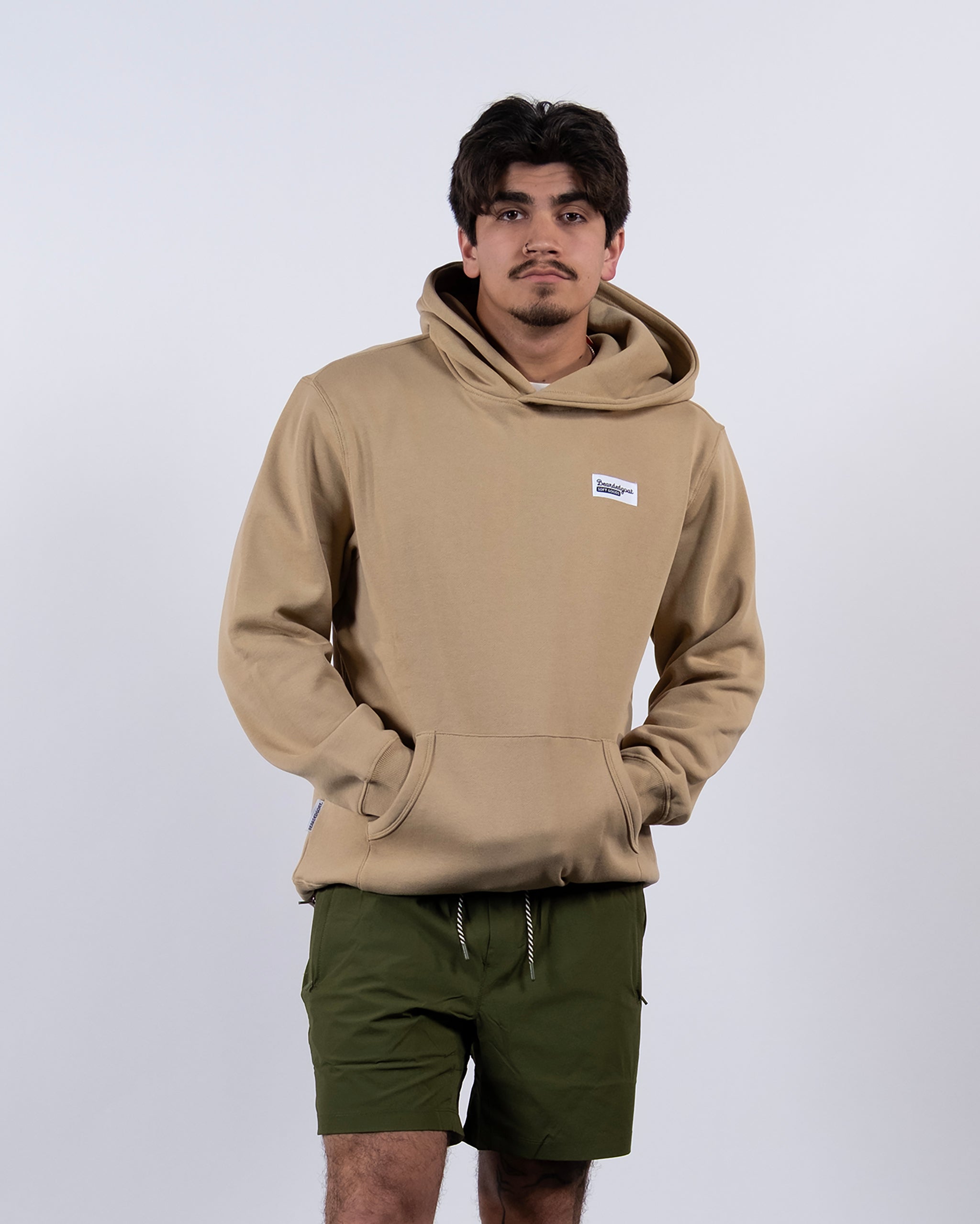 A person wears the tan beardedgoat The VOID™ Hoodie made from heavyweight cotton fleece with a front pocket, paired with olive green shorts, standing against a white background with hands in the hoodie pocket, looking at the camera.