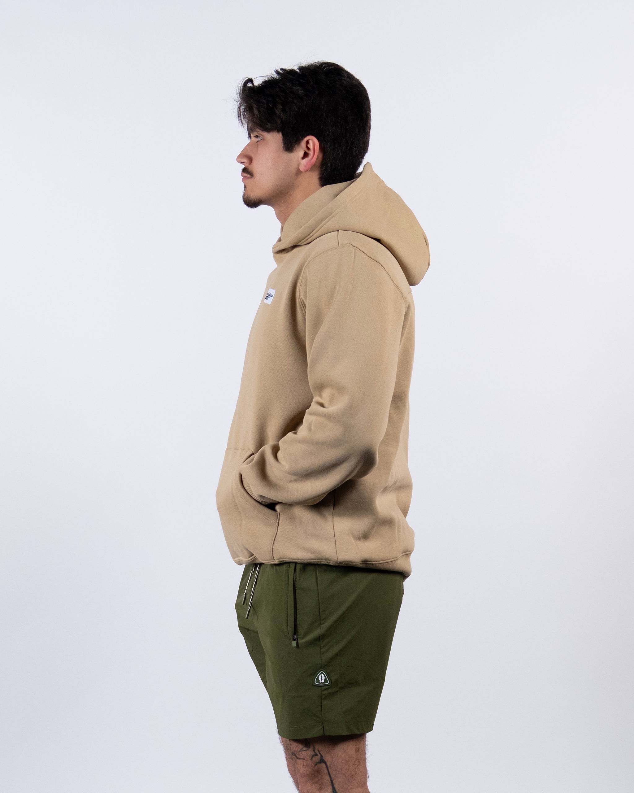 A man in profile stands against a plain white background, wearing the beardedgoat VOID™ Hoodie in tan—made from premium heavyweight cotton fleece—and olive green shorts, with his hands tucked into the hoodie’s front pocket.