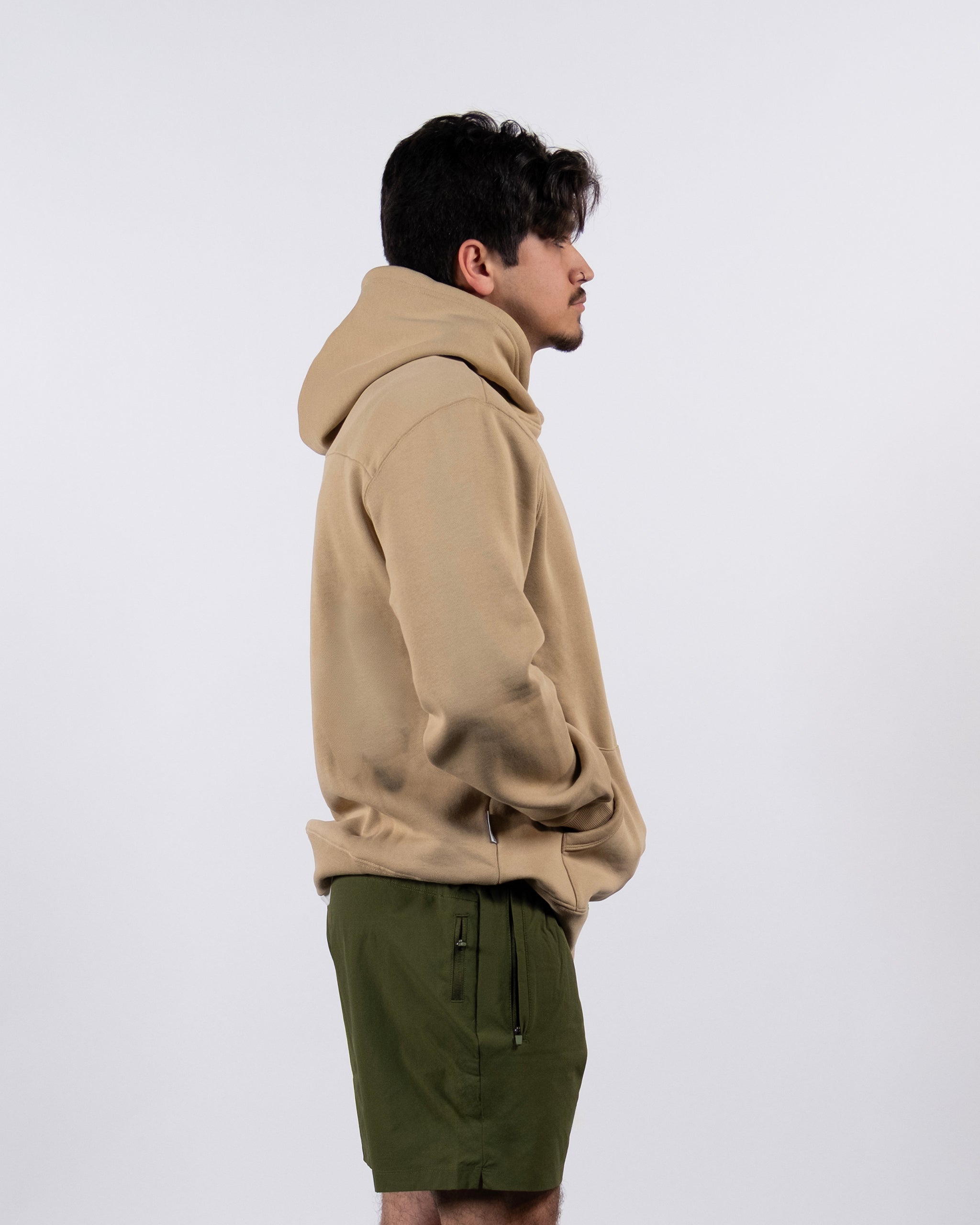 A person stands in profile against a white background, wearing the beardedgoat The VOID™ Hoodie with the hood down and olive green shorts with zippered pockets, hands in their hoodie pockets.
