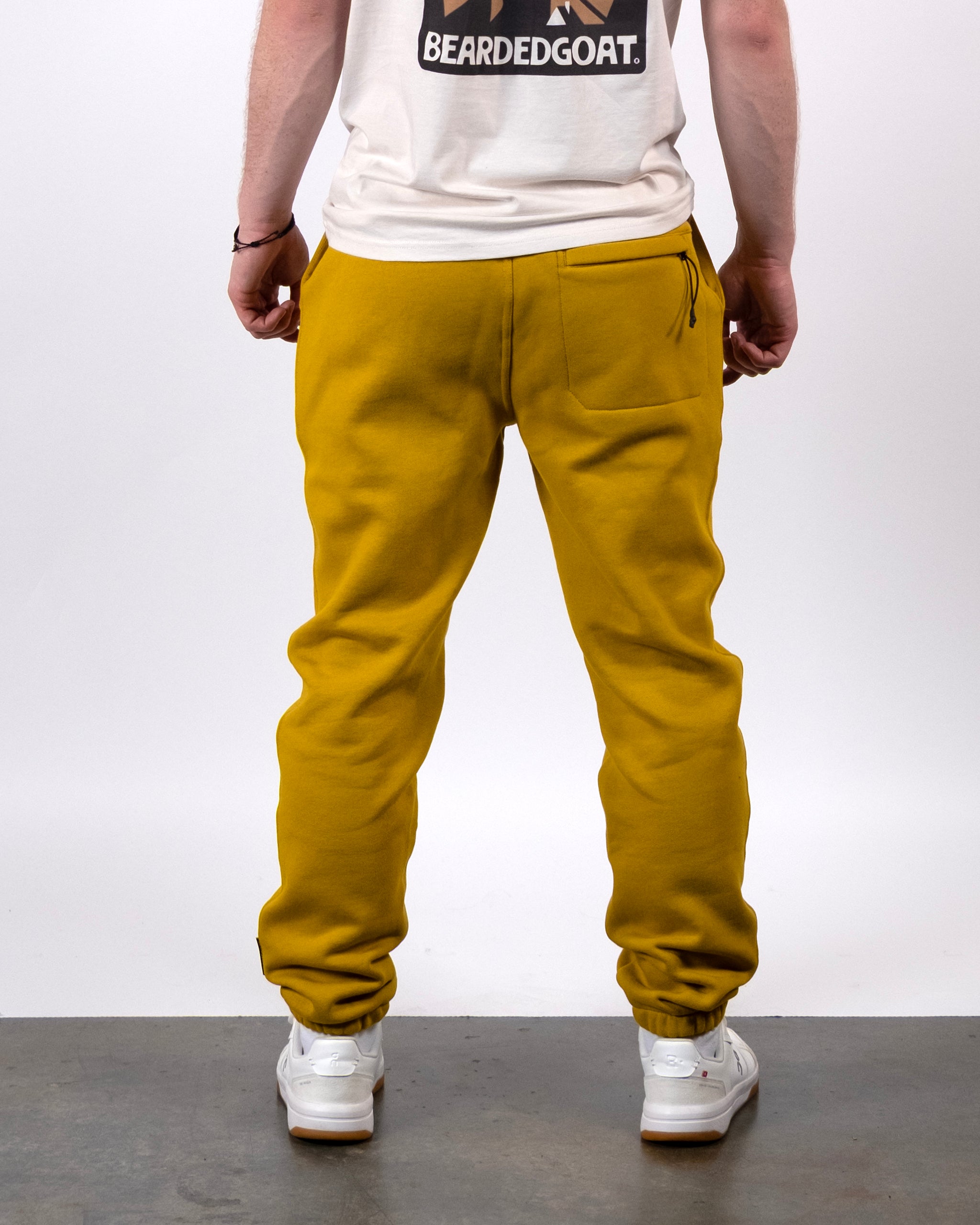 A person stands facing away, wearing a white beardedgoat t-shirt, mustard yellow VOID™ Sweatpant with a back pocket, and white sneakers, against a plain background.