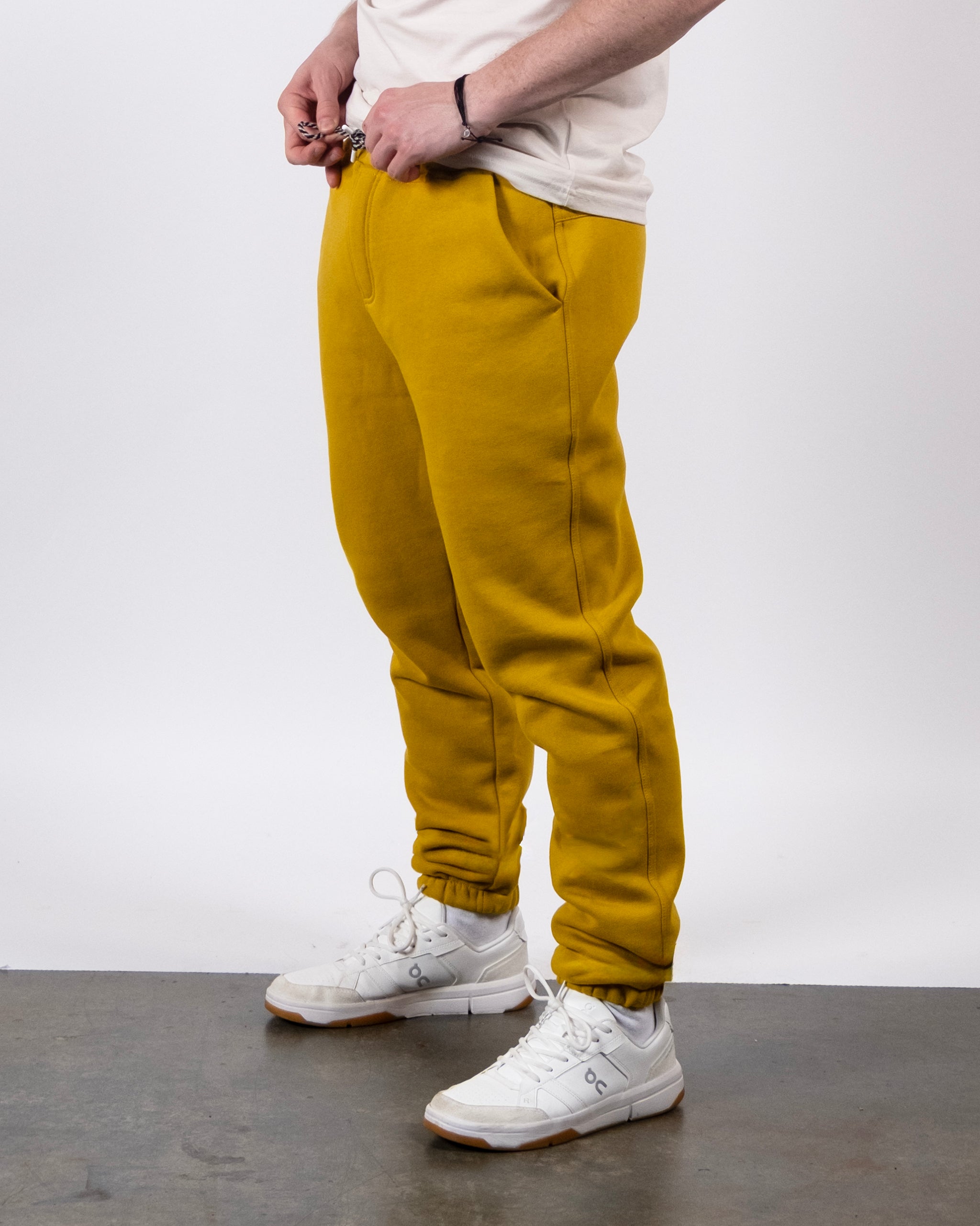 A person stands on a concrete floor wearing beardedgoat VOID™ Sweatpant in mustard yellow, a white T-shirt, and white sneakers. The cropped image shows the lower torso and legs, with hands adjusting the sweatpant waistband.