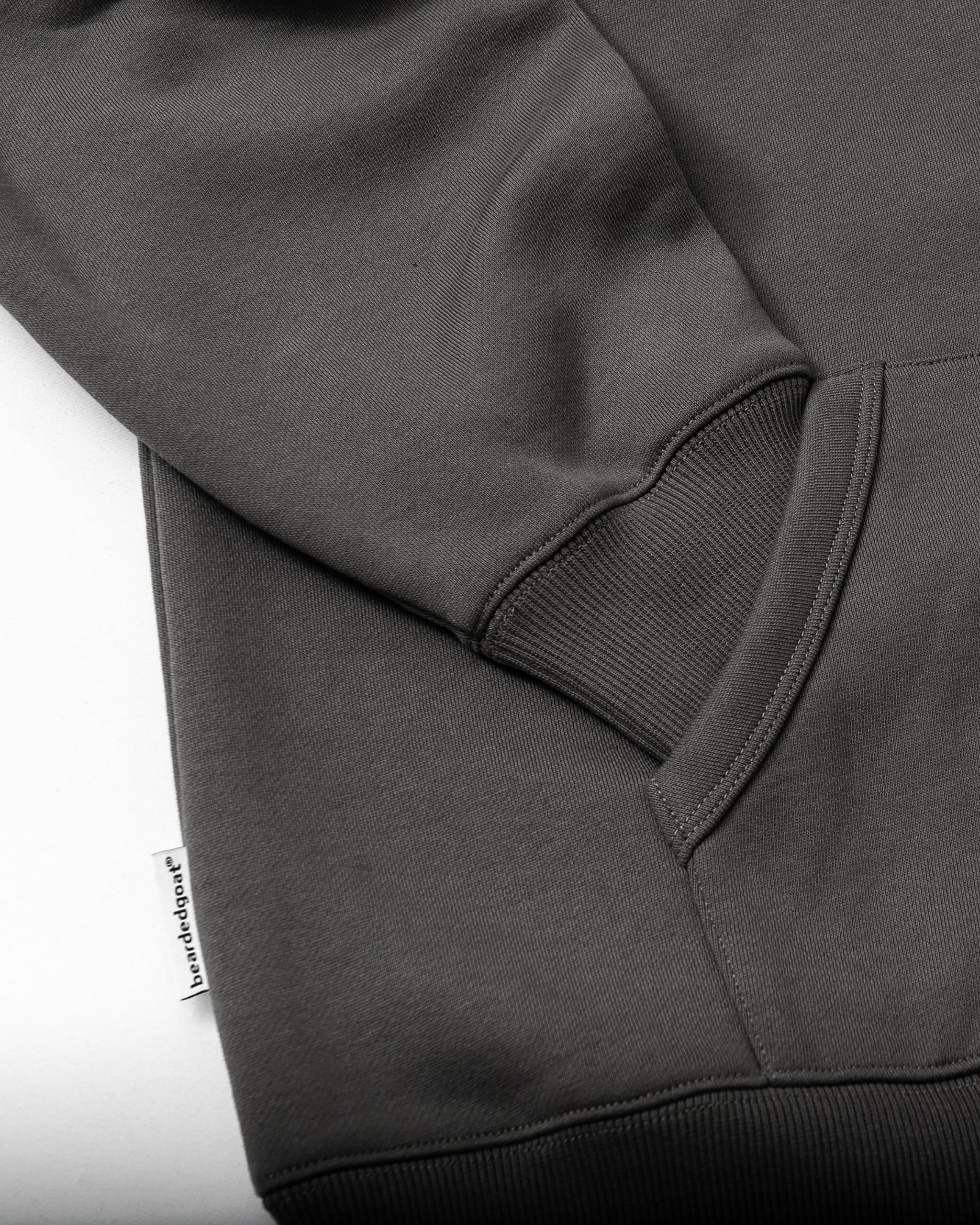 A close-up of the beardedgoat Men's Void Hoodie in dark gray heavyweight cotton highlights the ribbed cuff and hem, kangaroo pocket, and a small white fabric tag with text by the seam on a white background.