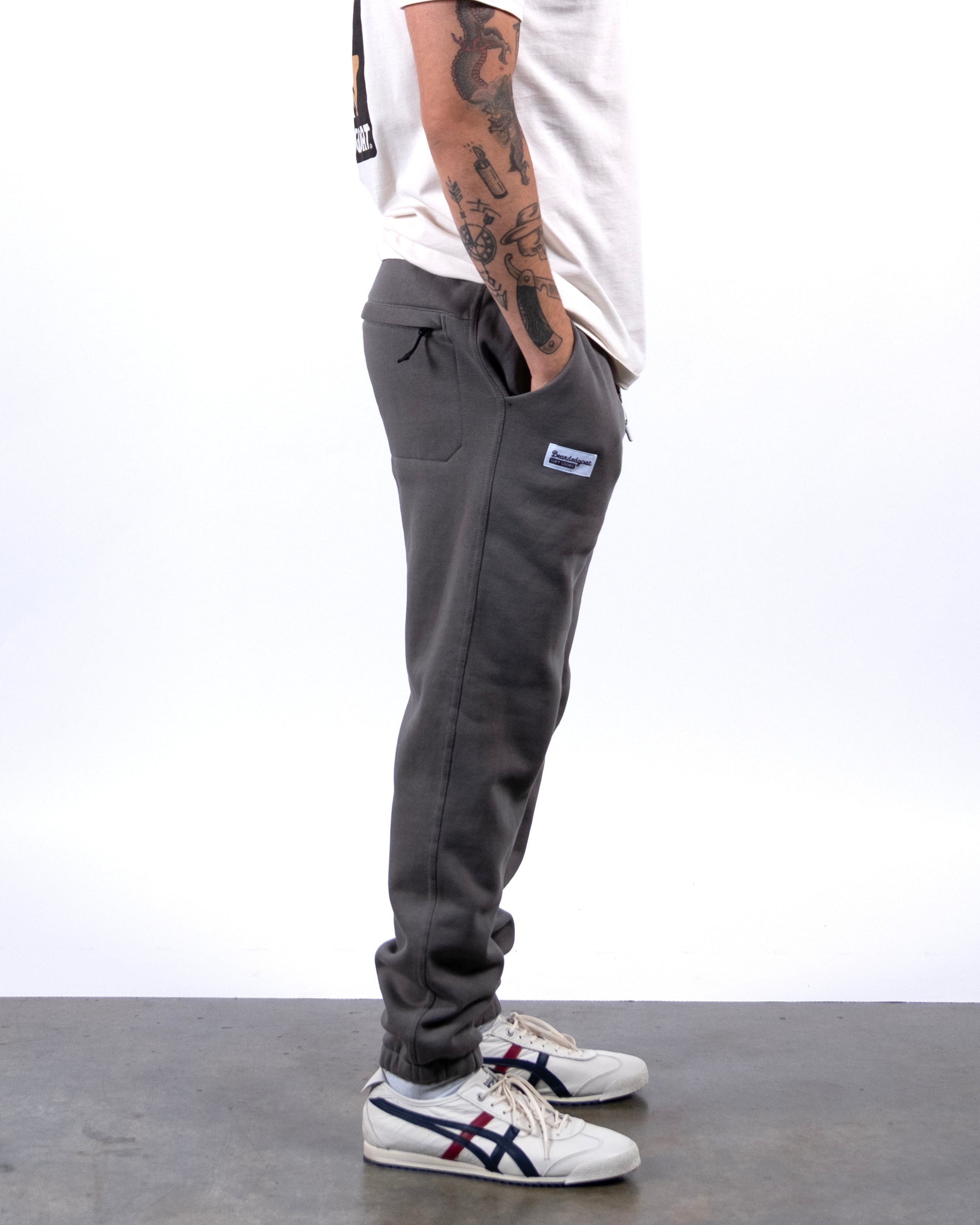 A person stands in profile wearing a white shirt, beardedgoat VOID™ Sweatpant in gray with a subtle patch, and white sneakers with blue and red stripes. Their hands are tucked into their pockets, showing tattooed arms.