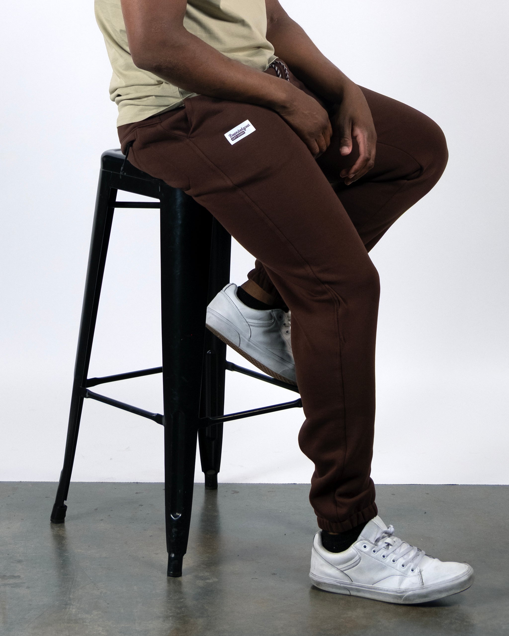 A person wearing beardedgoat VOID™ Sweatpants and white sneakers is seated on a black metal stool against a plain white background, with only their lower body shown.