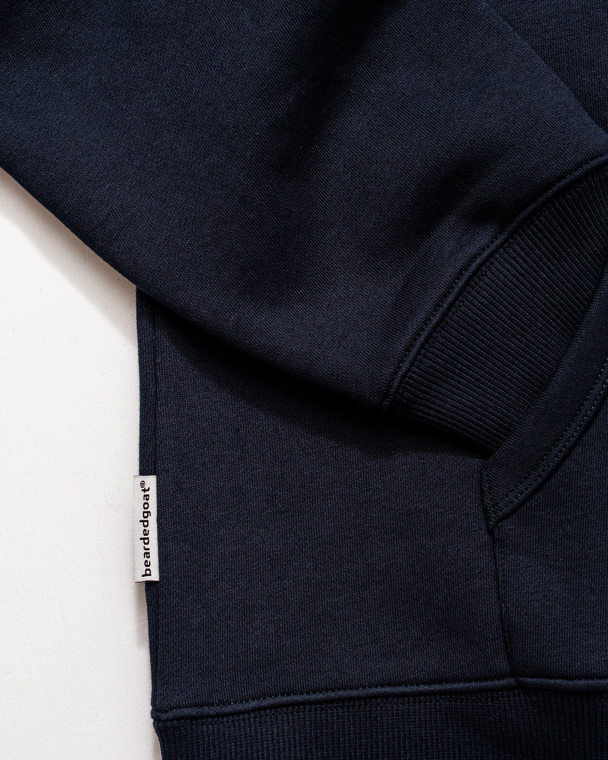 Close-up of a navy blue Men's Void Hoodie from beardedgoat, crafted from heavyweight cotton, highlighting the sleeve, ribbed cuff, and a small white side seam tag featuring the brand name.