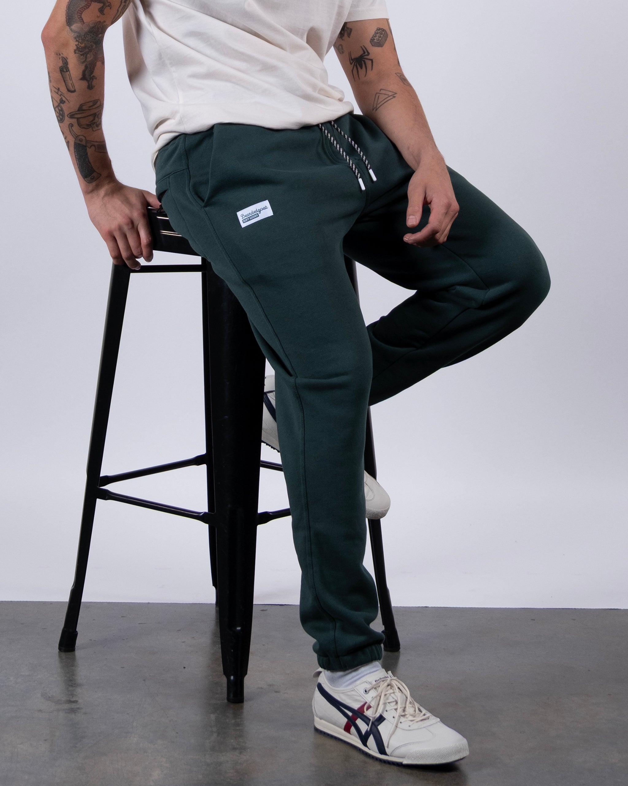 A person with tattooed arms sits on a tall black stool in a white t-shirt and beardedgoat VOID™ Sweatpant—dark green joggers with a patch—paired with white sneakers featuring blue and red stripes, set against a plain light background.