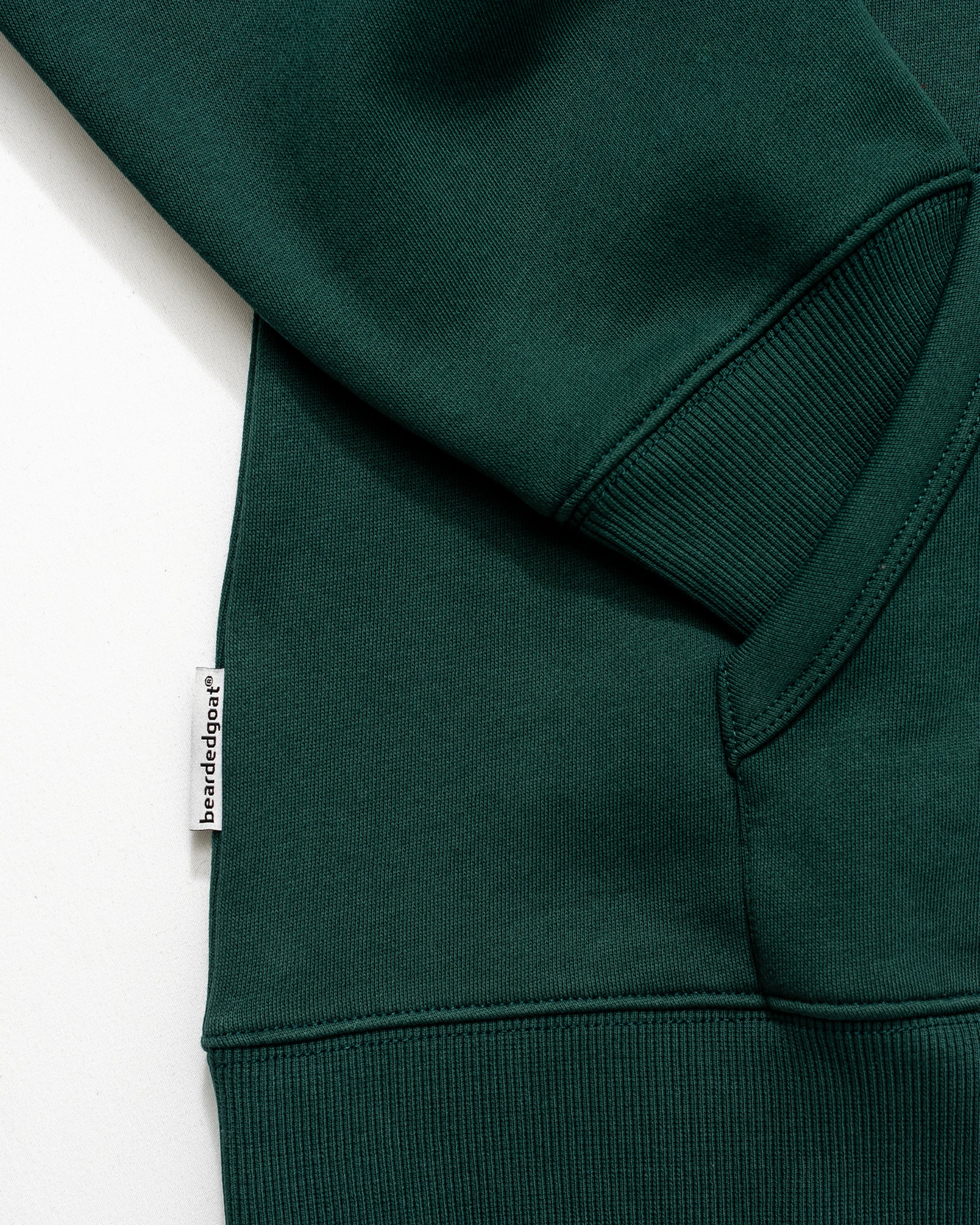 Close-up of the Men's Void Hoodie by beardedgoat in forest green heavyweight cotton, featuring a ribbed cuff and waistband, slanted pocket, and a white beardedgoat tag on the seam.
