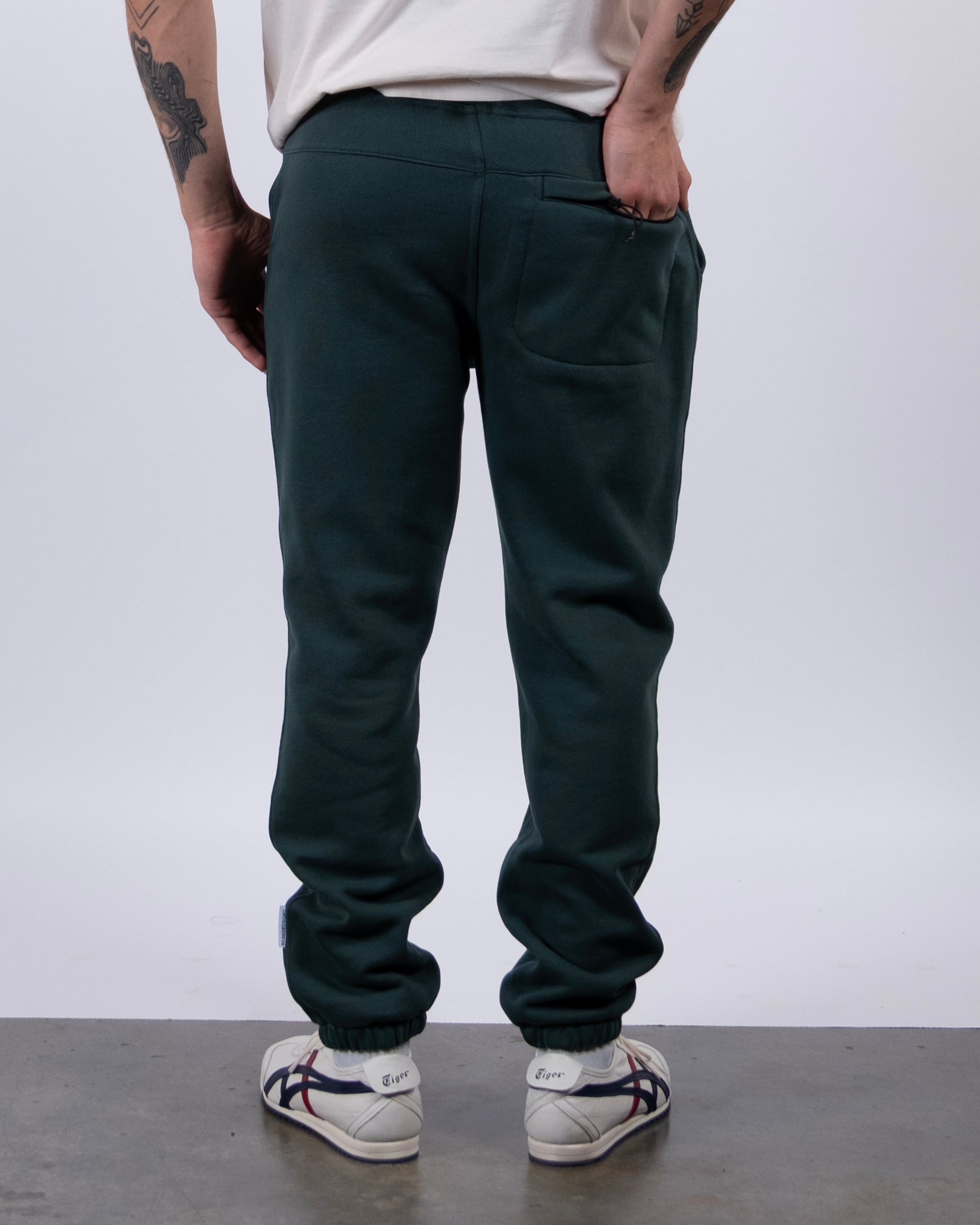 A person stands with their back to the camera, wearing beardedgoat VOID™ Sweatpant in dark green, a white shirt, and white sneakers with dark stripes. Tattoos are visible on their right arm. The background is plain gray.