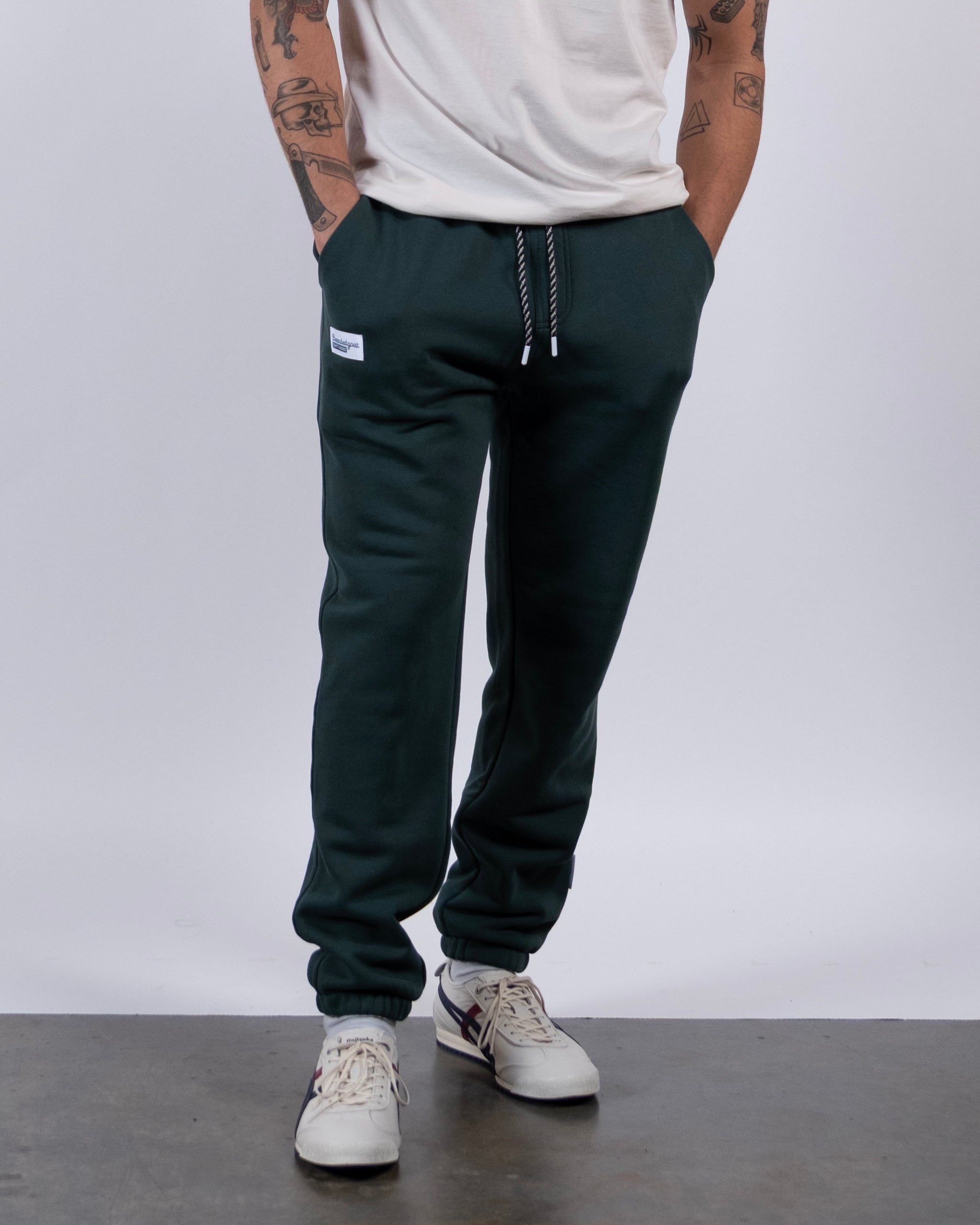A person wears the beardedgoat VOID™ Sweatpant in dark green with a drawstring, paired with a white t-shirt and white sneakers. Standing against a plain background, their tattooed arms are visible as they keep their hands in their pockets.