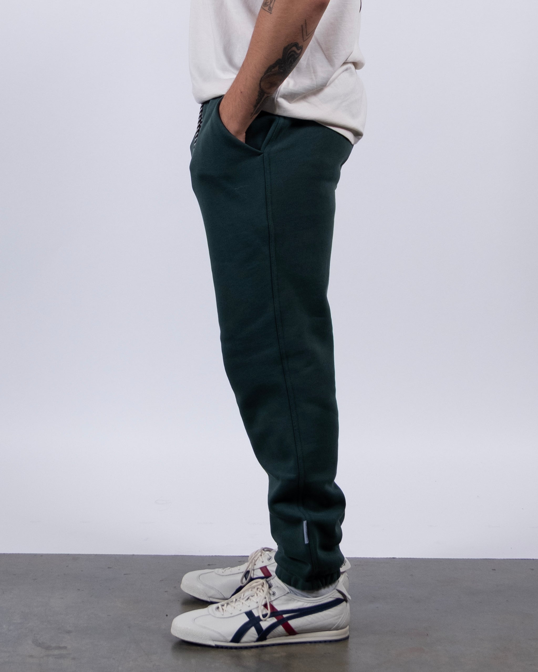 A person stands in profile with hands in pockets, wearing a white shirt and beardedgoat VOID™ Sweatpant in dark green, paired with white sneakers with blue and red stripes, against a plain light gray background.