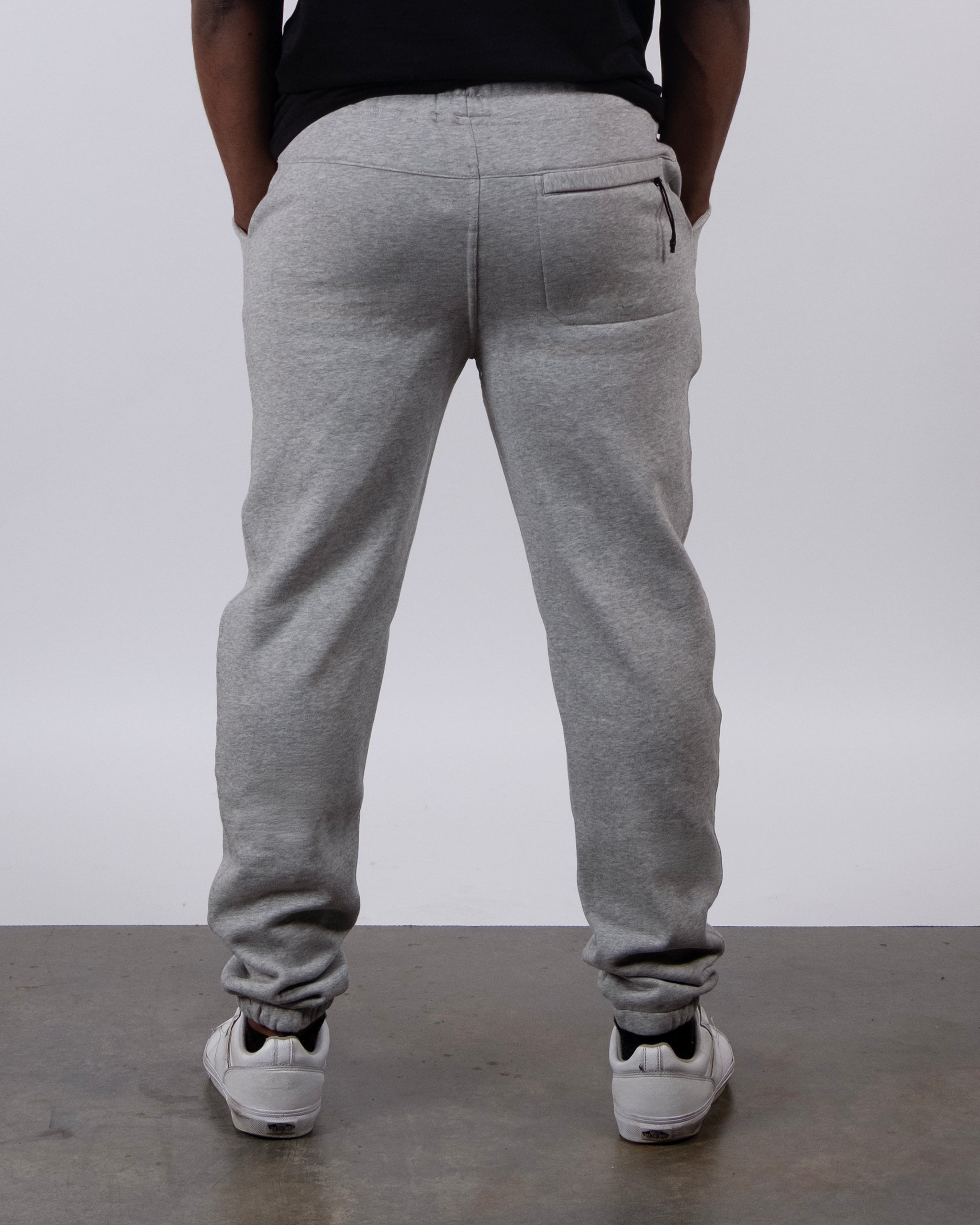 A person faces away from the camera, hands in pockets, wearing beardedgoat VOID™ Sweatpant in light gray with a black shirt and white sneakers. The plain, neutral background highlights the premium men's sweats.