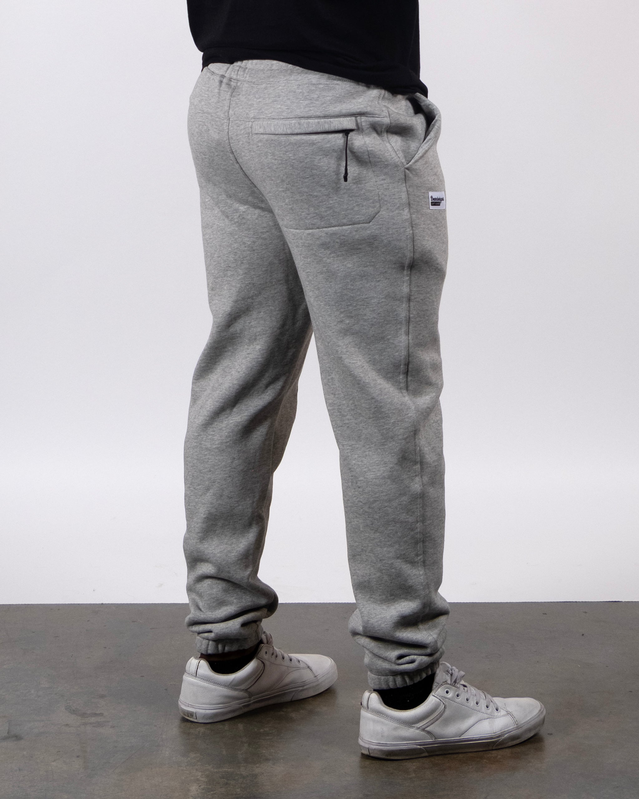A person in beardedgoat VOID™ Sweatpant—light gray heavyweight fleece—paired with white sneakers and a black shirt stands on a concrete floor, shown from the side and back against a plain white background.