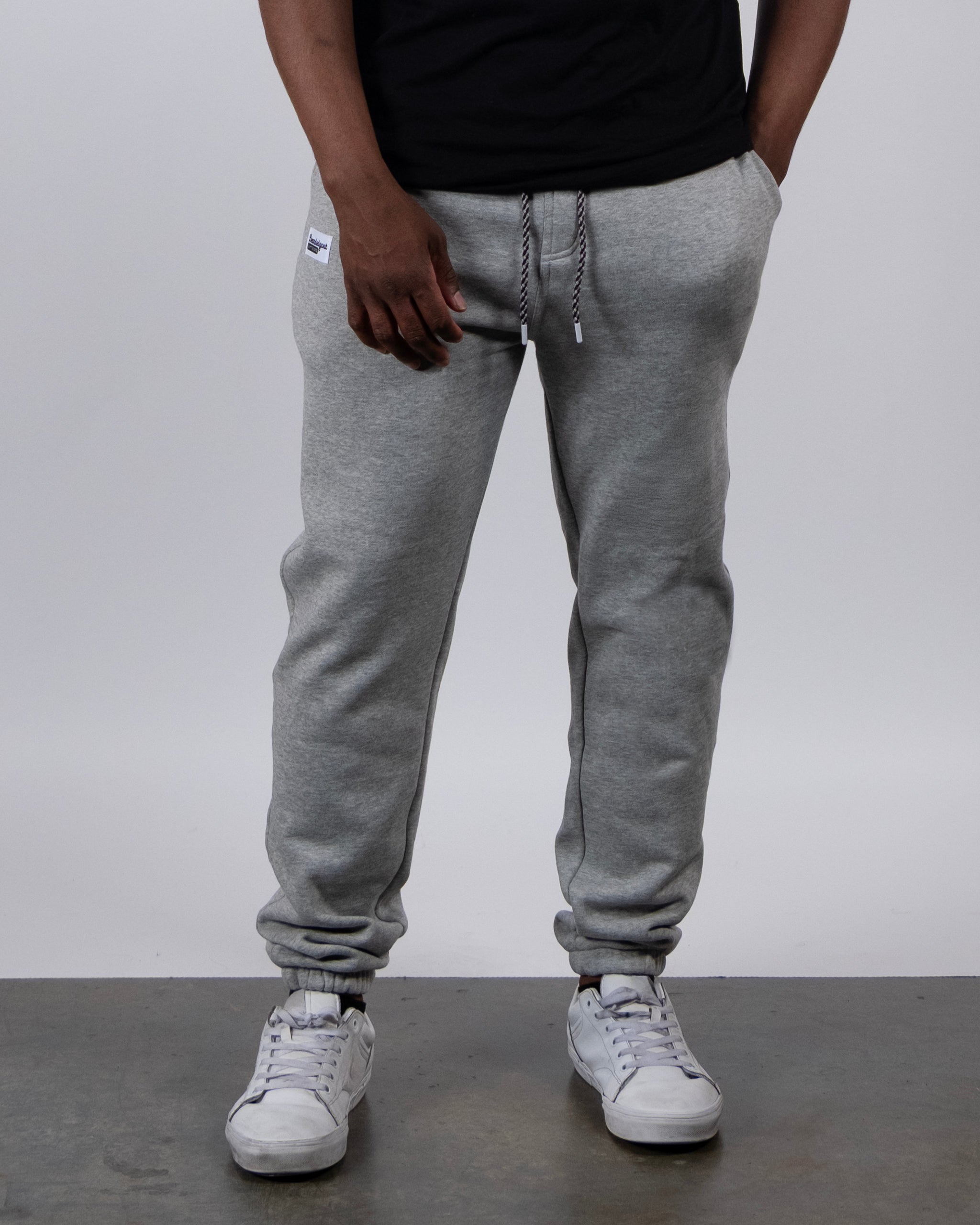 A person wears beardedgoat’s VOID™ Sweatpant in light gray, paired with a black t-shirt and white sneakers. They stand on a concrete floor against a plain white background, arms relaxed at their sides.