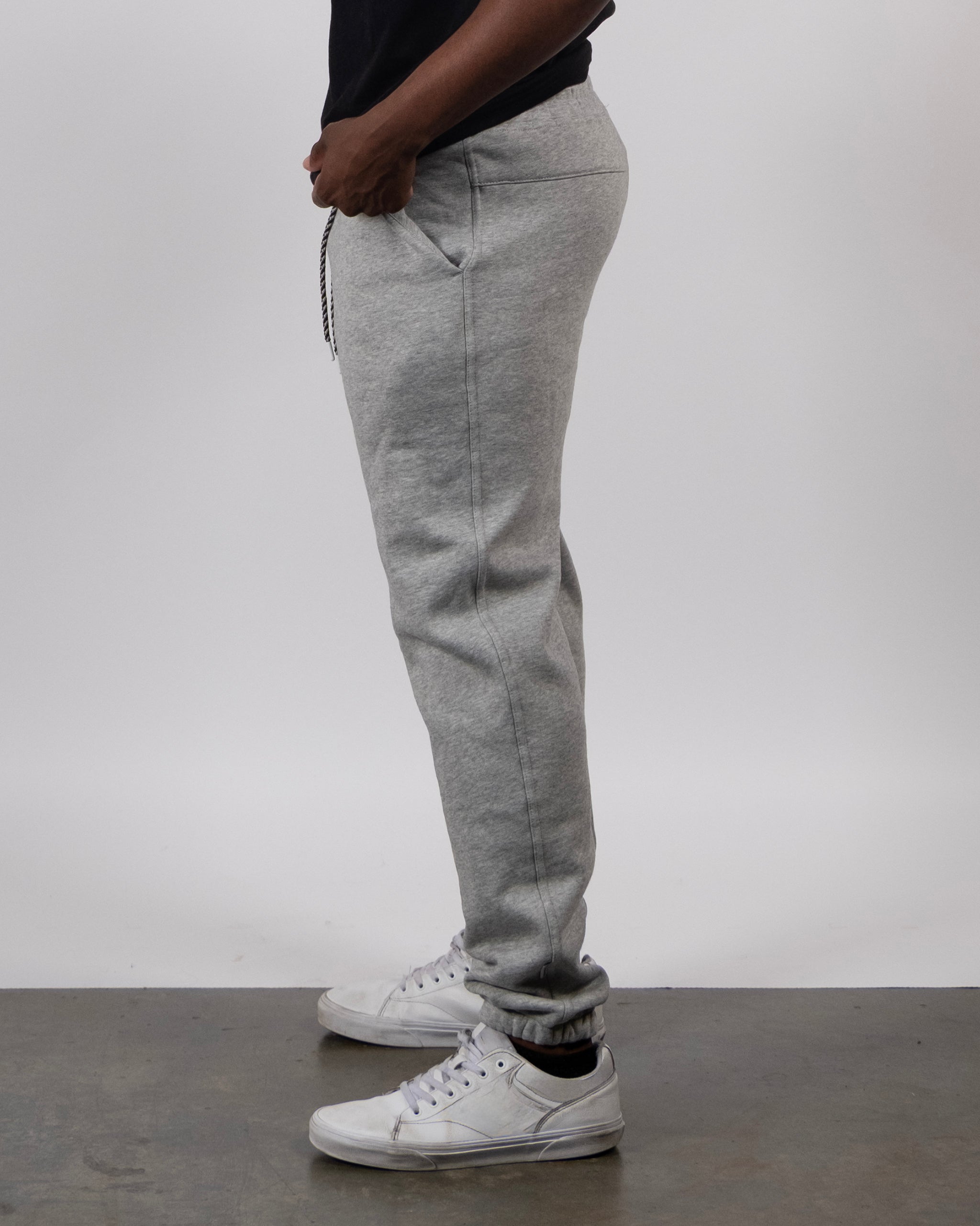 A person in a black shirt, beardedgoat VOID™ Sweatpant in light gray, and white sneakers stands sideways against a plain light background with hands in their pocket.