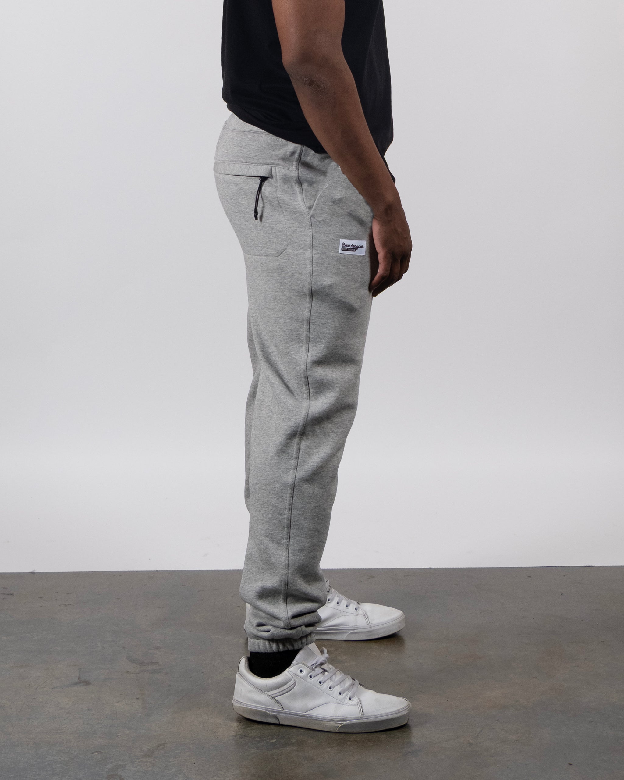 A person stands sideways on a concrete floor, wearing a black shirt, beardedgoat VOID™ Sweatpant in light gray heavyweight fleece with pockets, and white sneakers. The background is plain and light-colored.