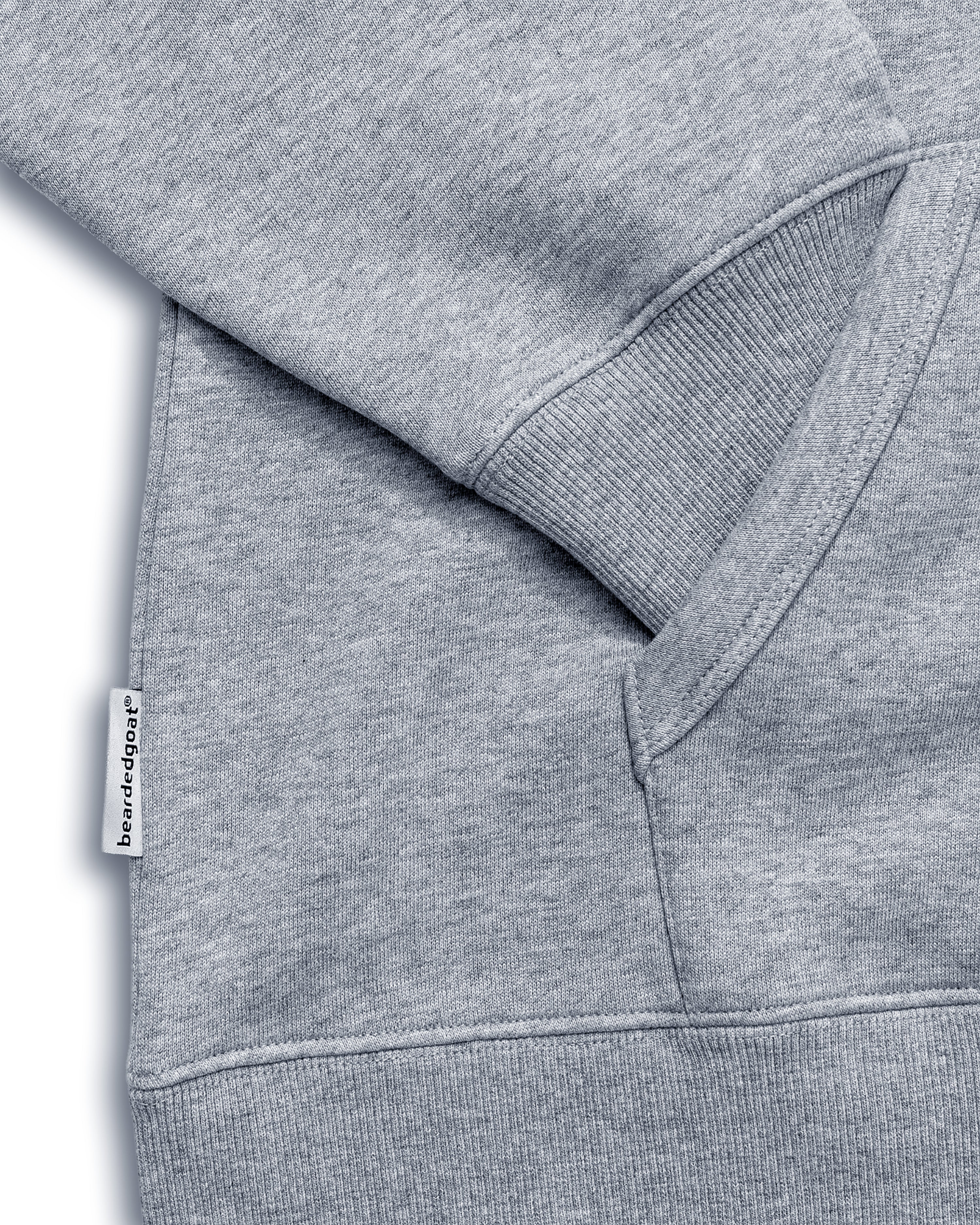 Close-up of the VOID™ Hoodie by beardedgoat in heather gray, made from heavyweight cotton fleece. Shows a ribbed cuff, pocket seam, and a small white “beardedgoat” tag. Fabric texture and stitching details are visible.
