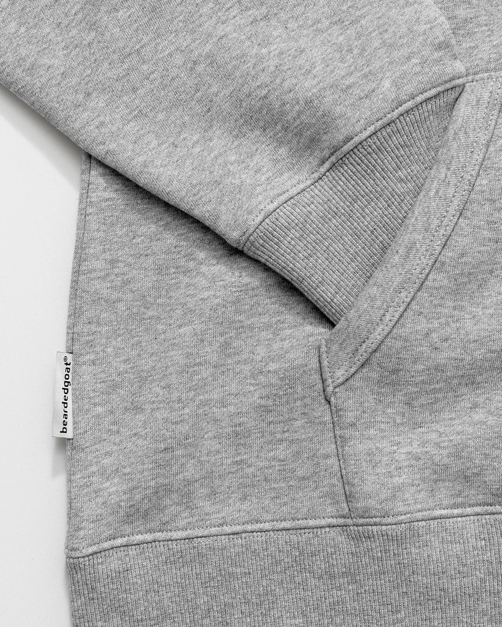 A close-up of The VOID™ Hoodie by beardedgoat in light gray shows its ribbed cuff, pocket seam, and a small white tag with black text. The heavyweight cotton fleece fabric looks soft with a subtle texture.
