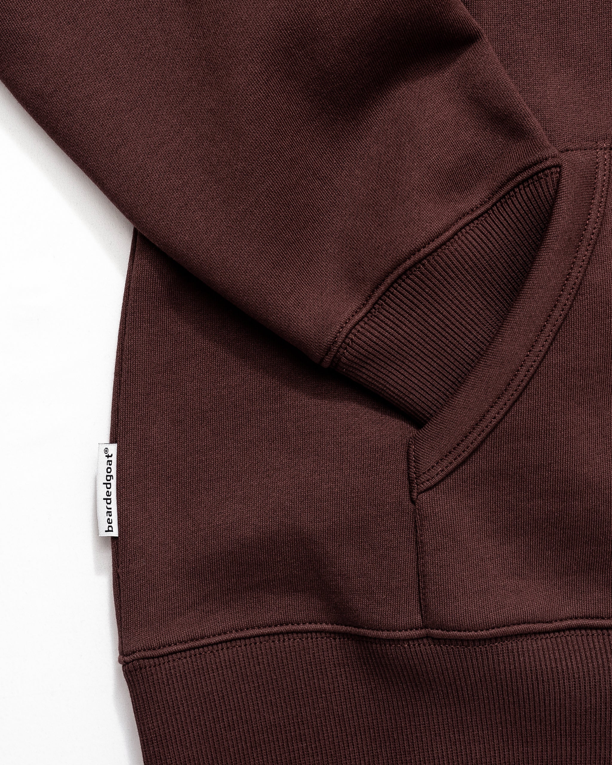 Close-up of the Men's Void Hoodie by beardedgoat in brown, featuring a ribbed cuff, partial pocket, and a small white "besidestaget" tag on the side seam—made from heavyweight cotton for lasting durability.
