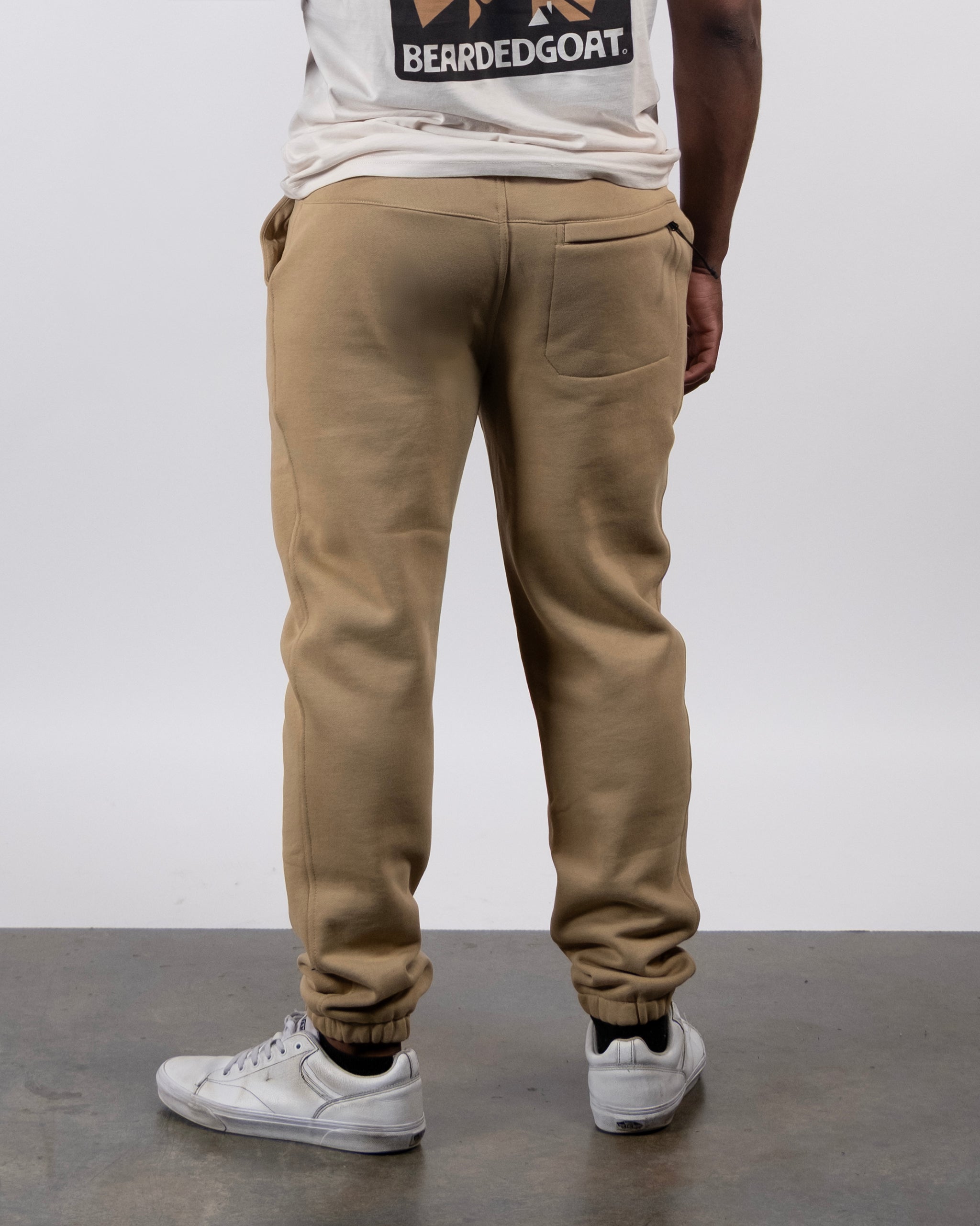 A person stands facing away, wearing beardedgoat’s VOID™ Sweatpant, a white T-shirt with a visible beardedgoat logo on the back, and white sneakers against a plain background.