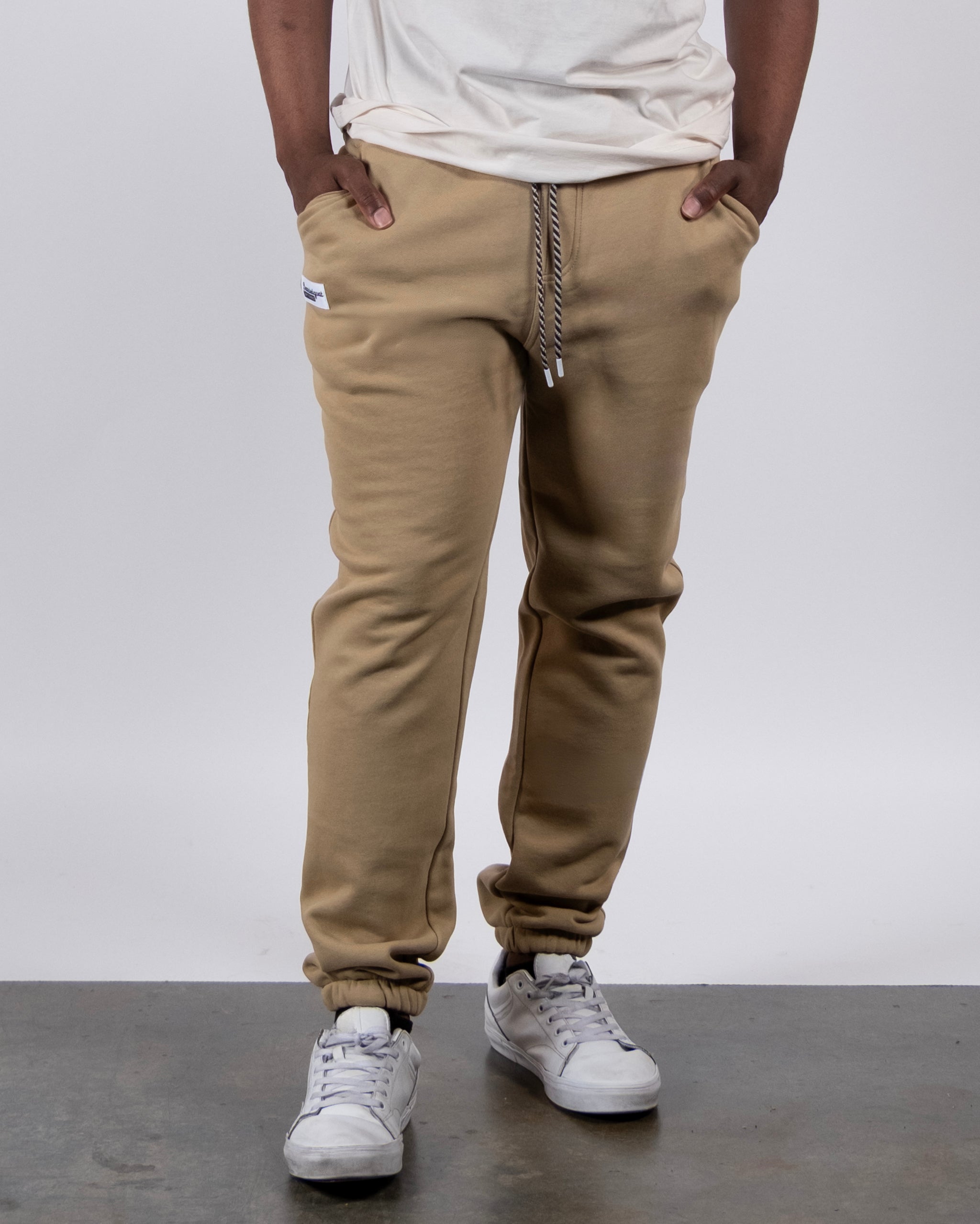 A person wearing a white T-shirt, VOID™ Sweatpant tan joggers by beardedgoat with a drawstring, and white sneakers stands with hands in their pockets against a plain white background.