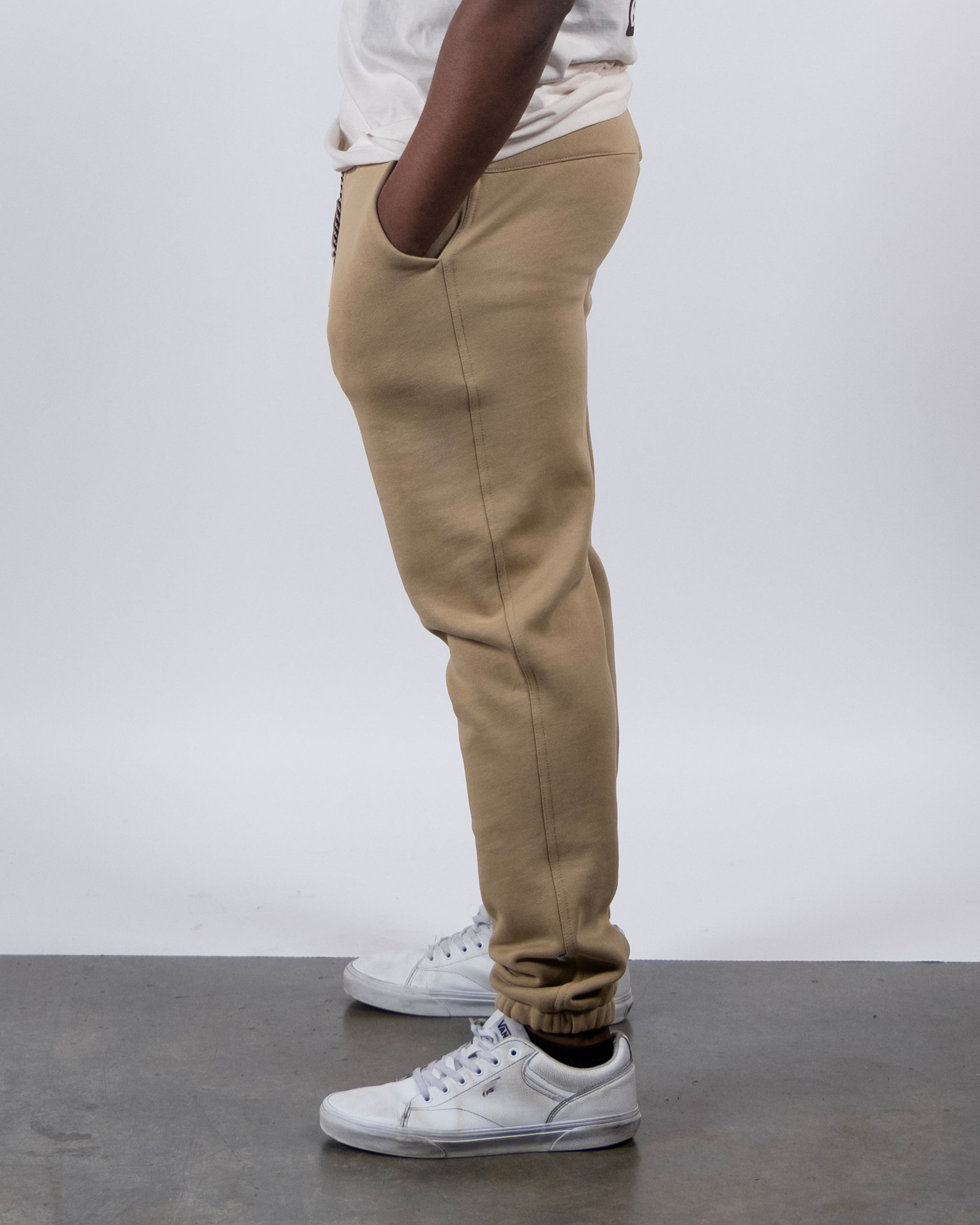 A person stands sideways with hands in pockets, wearing beardedgoat VOID™ Sweatpant in tan jogger style, paired with a white t-shirt and white sneakers against a plain light gray background.