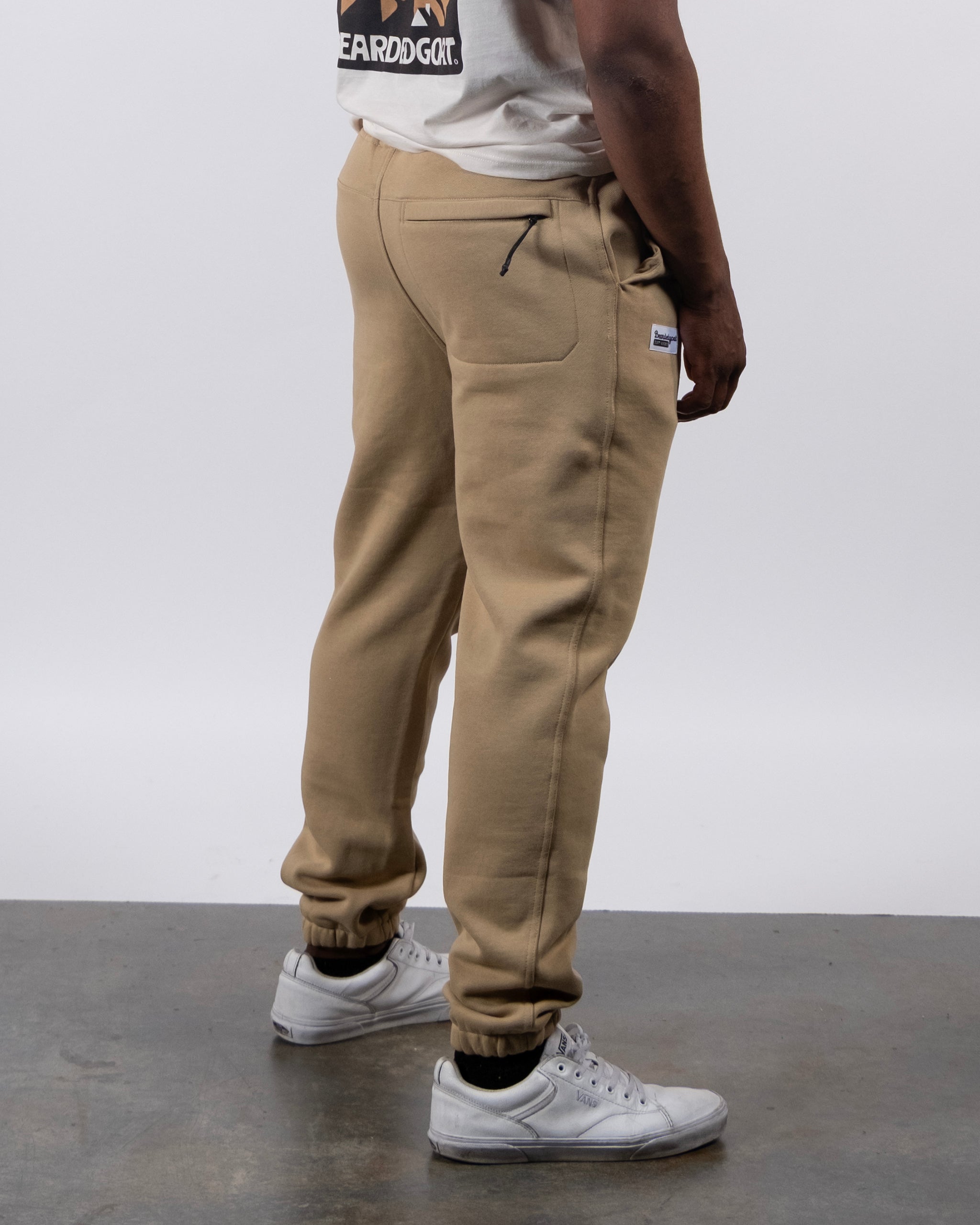 Someone wearing beardedgoat VOID™ Sweatpants in tan, a white T-shirt, and white sneakers stands on a concrete floor against a plain light background, facing slightly away from the camera.