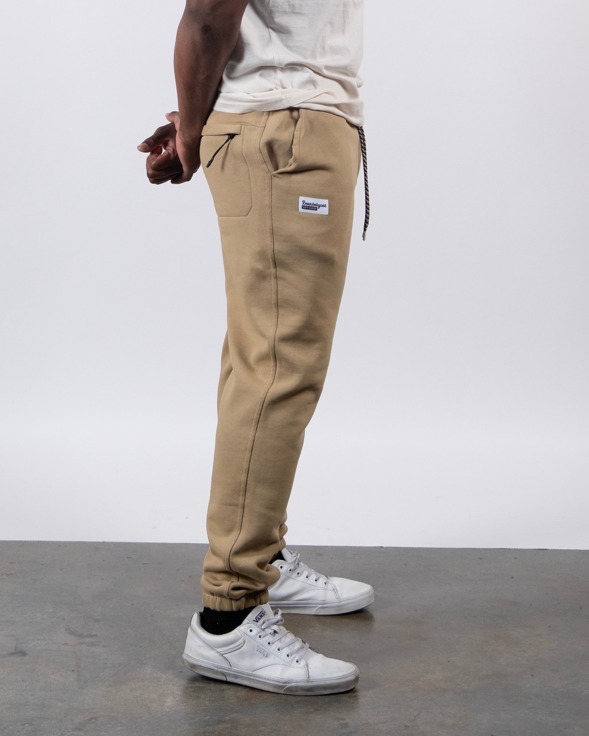A person in a white shirt and beardedgoat VOID™ Sweatpant stands sideways on a gray floor, hands behind back. The tan sweatpants feature a small rectangular hip label, drawstring waist, and are paired with white sneakers.