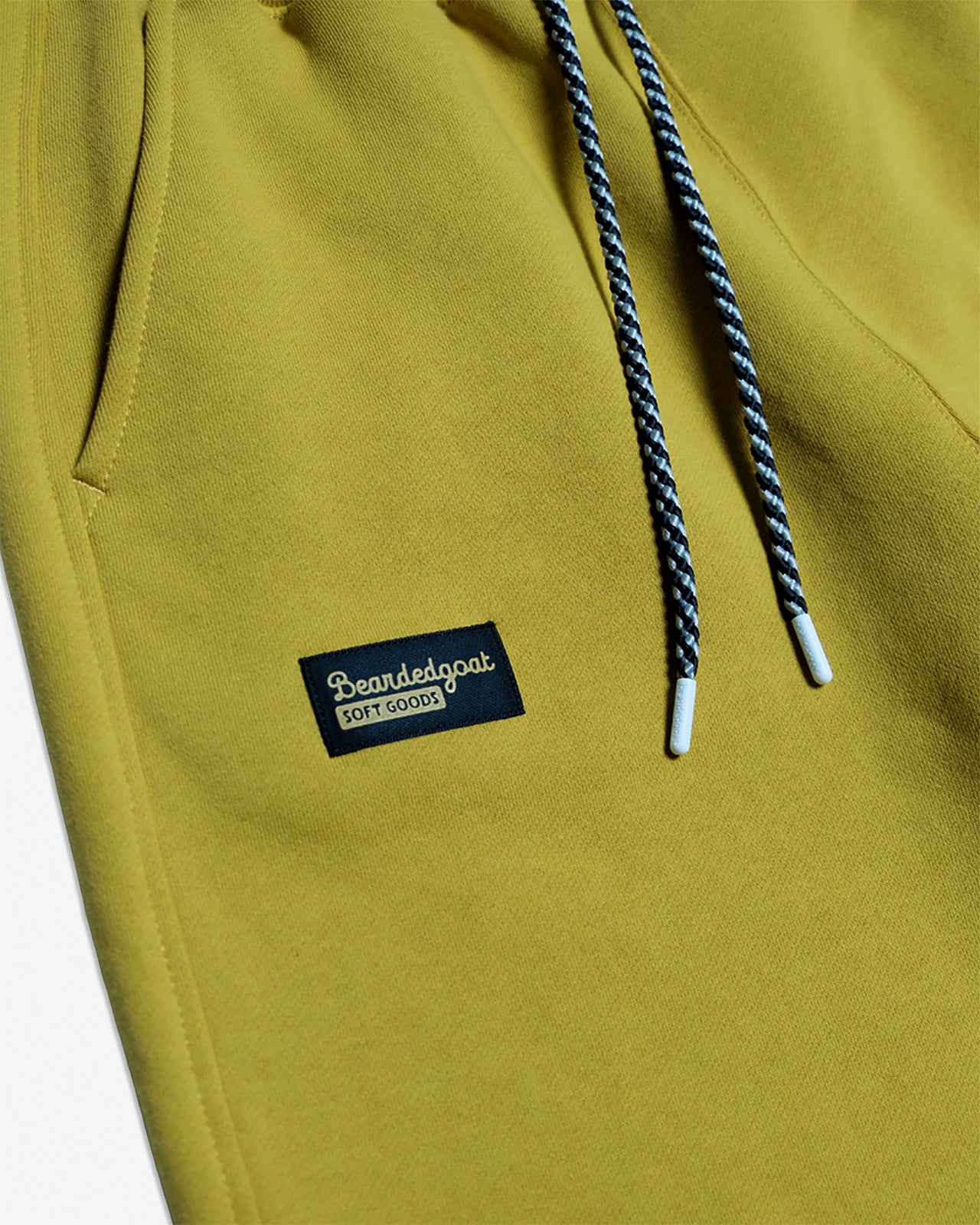 Close-up of yellow beardedgoat VOID™ Sweatpant featuring black and white drawstrings and a small black rectangular “beardedgoat Soft Goods” patch near the pocket.