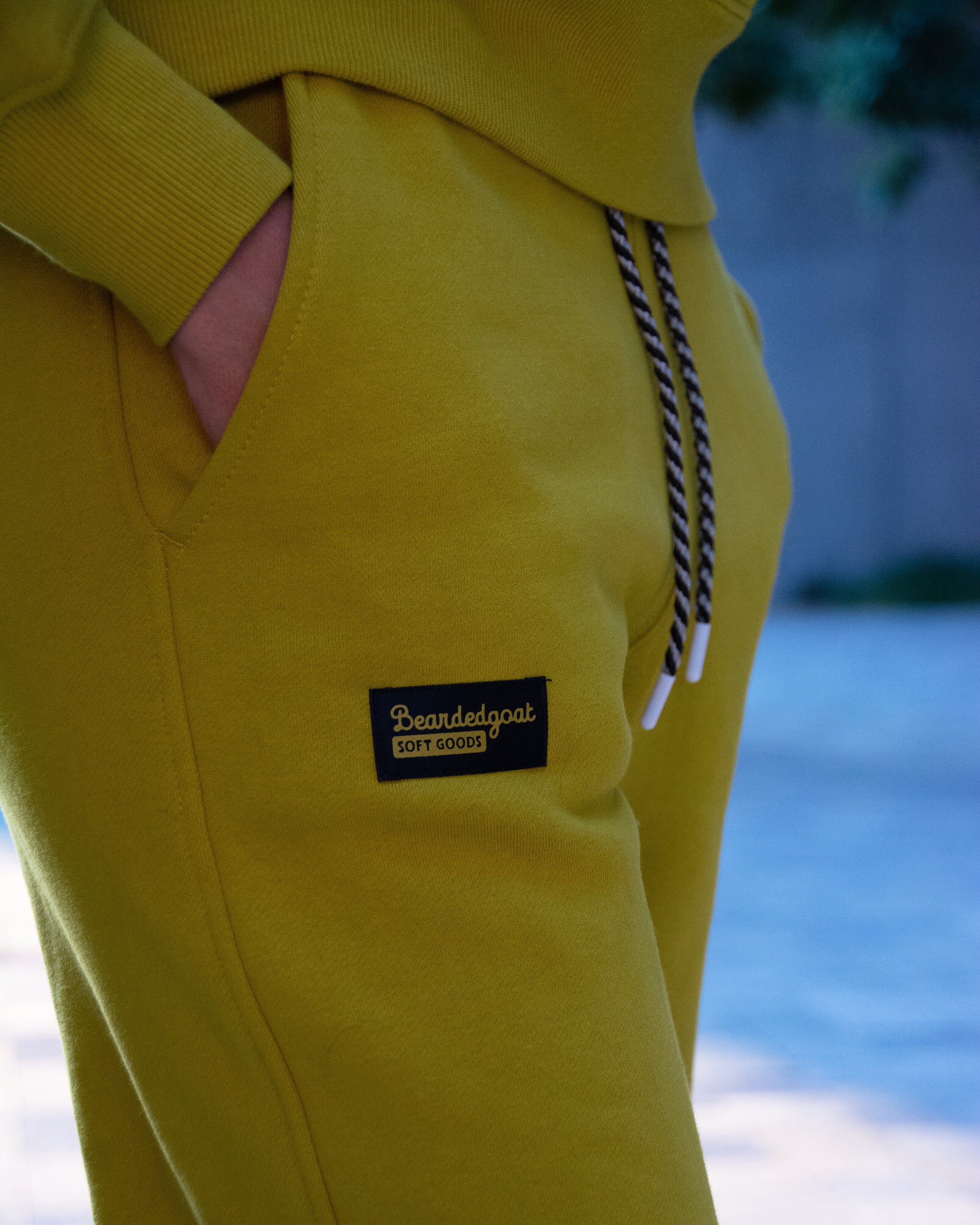 Close-up of a person wearing the NEW — Overnight™ Classic Jogger by BEARDEDGOAT in mustard yellow, featuring a black and white drawstring, “Beardedgoat Soft Goods” patch on the pocket, heavyweight cotton fleece, and a hand partially in the pocket.