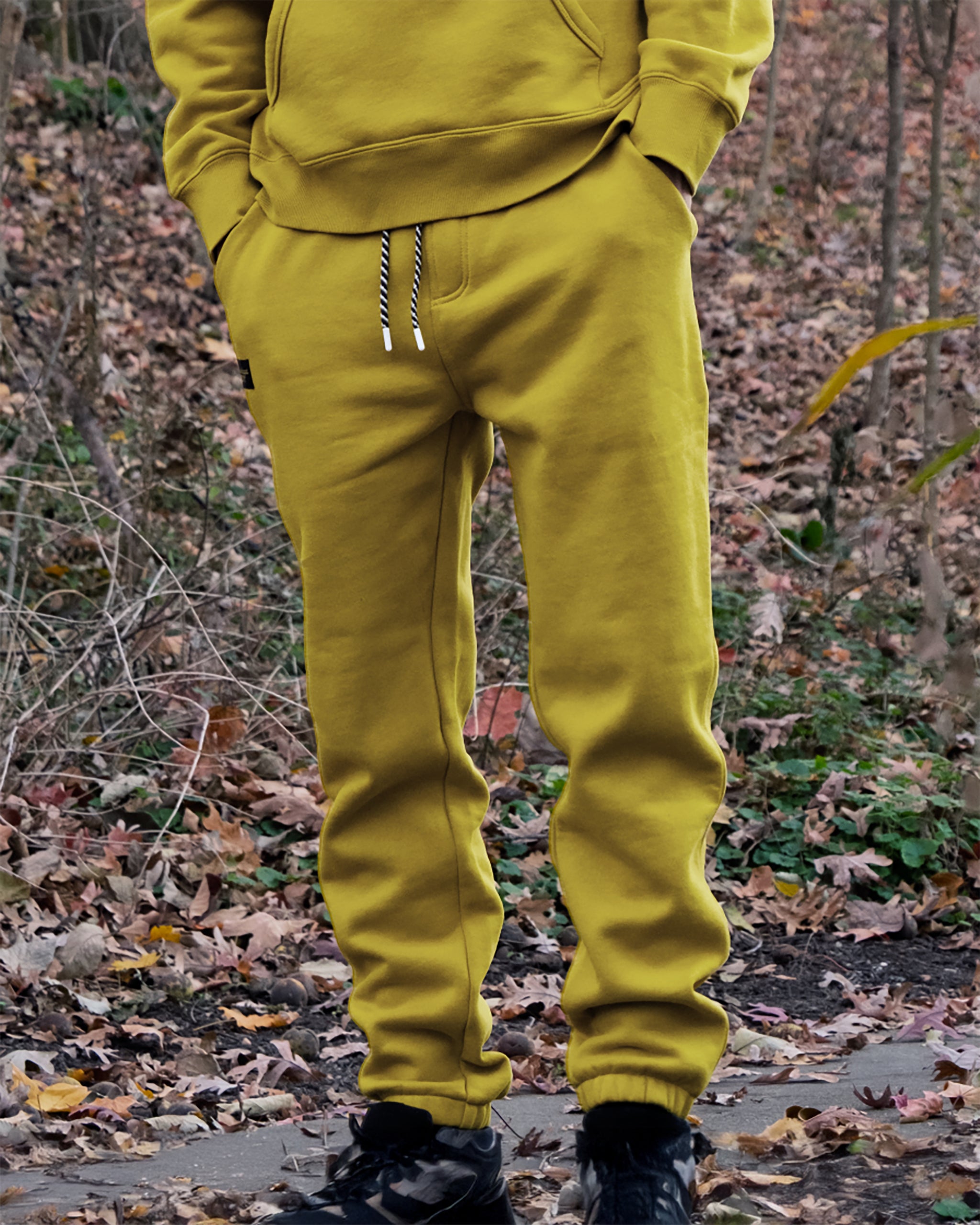 Outdoors on a leafy path, someone stands with hands in pockets wearing beardedgoat’s VOID™ Sweatpant and a mustard yellow hoodie, surrounded by trees and dry foliage.