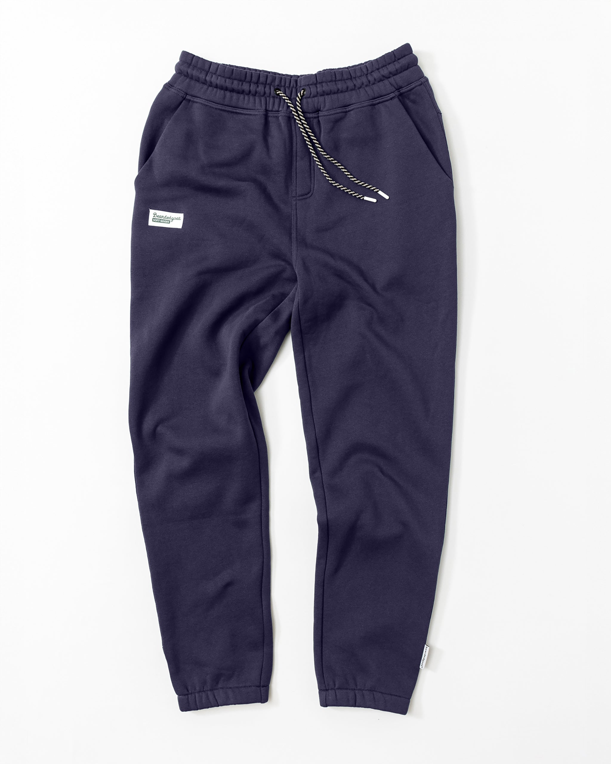 A pair of Men's Void Sweatpants by beardedgoat in dark navy blue, featuring an elastic waistband, drawstring, side pockets, a zippered back pocket, and a small white rectangular patch on the left thigh, laid flat on a white background.