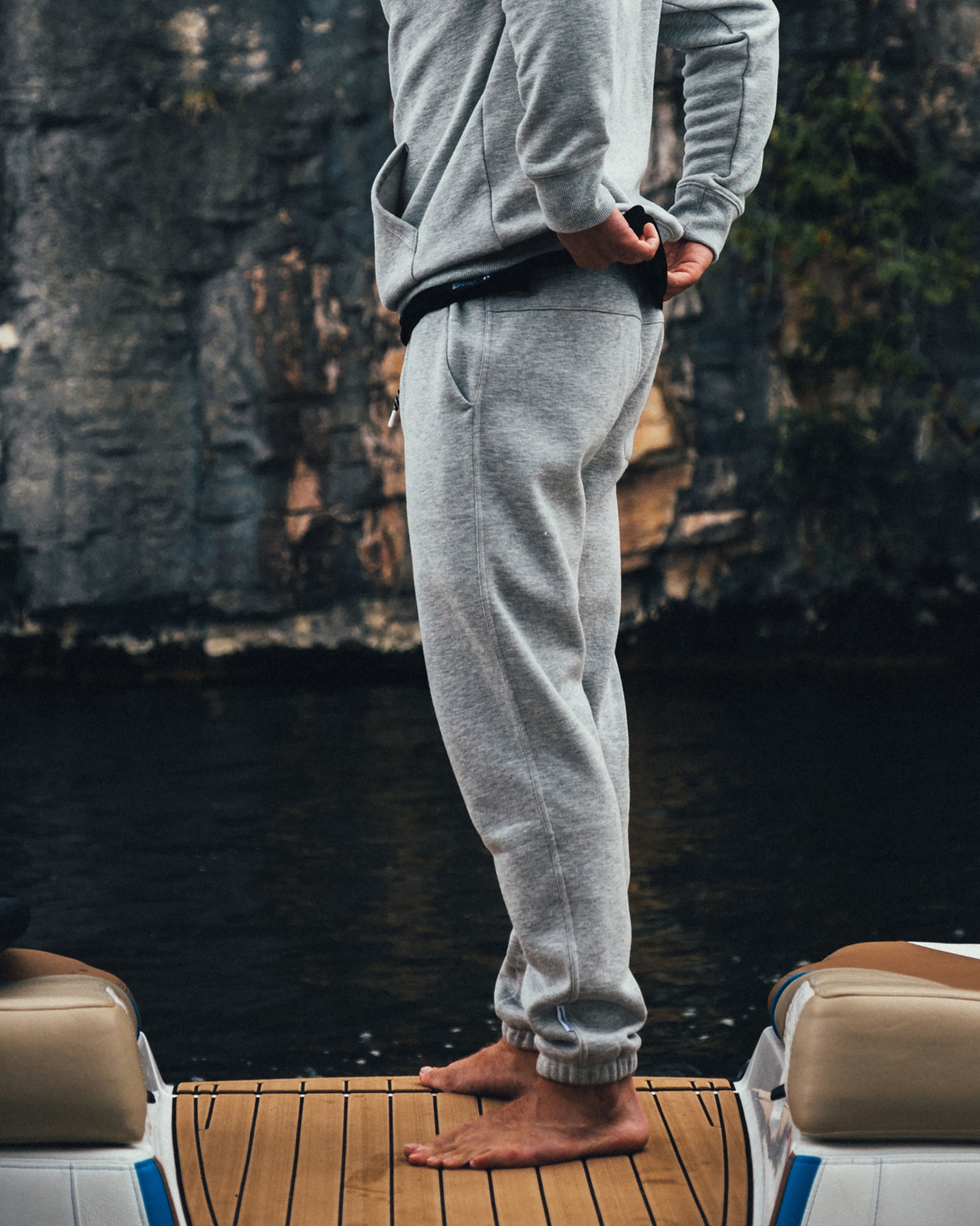 A person in beardedgoat Men's Void Sweatpants stands barefoot on a wooden dock, facing sideways with hands in pockets. The photo shows their body from neck to feet, with dark water and a rocky cliff in the background.