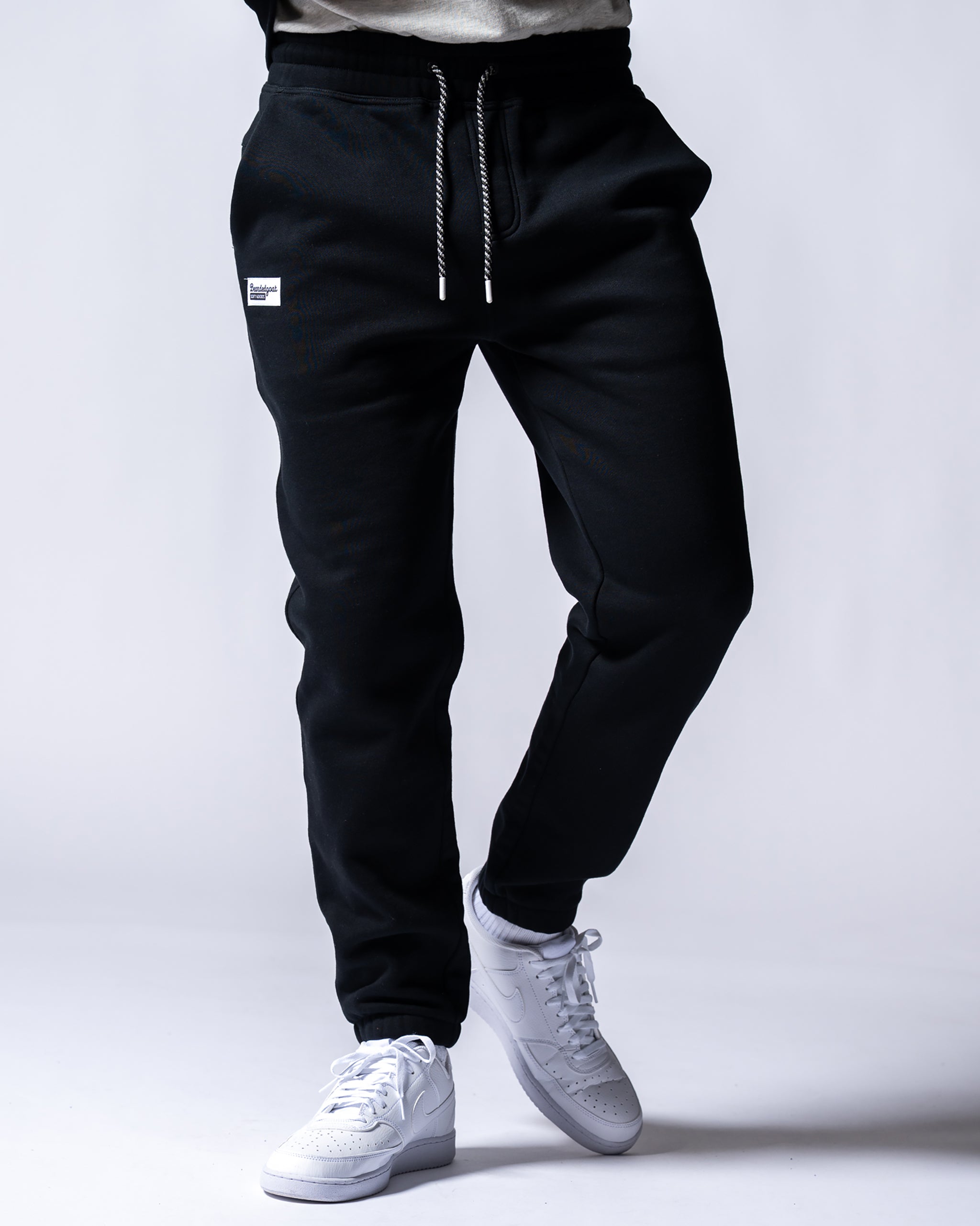 A person wears beardedgoat Men's Void Sweatpants, black joggers with a drawstring and zipper back pocket, paired with white sneakers, standing against a plain light background. The upper body is not visible.