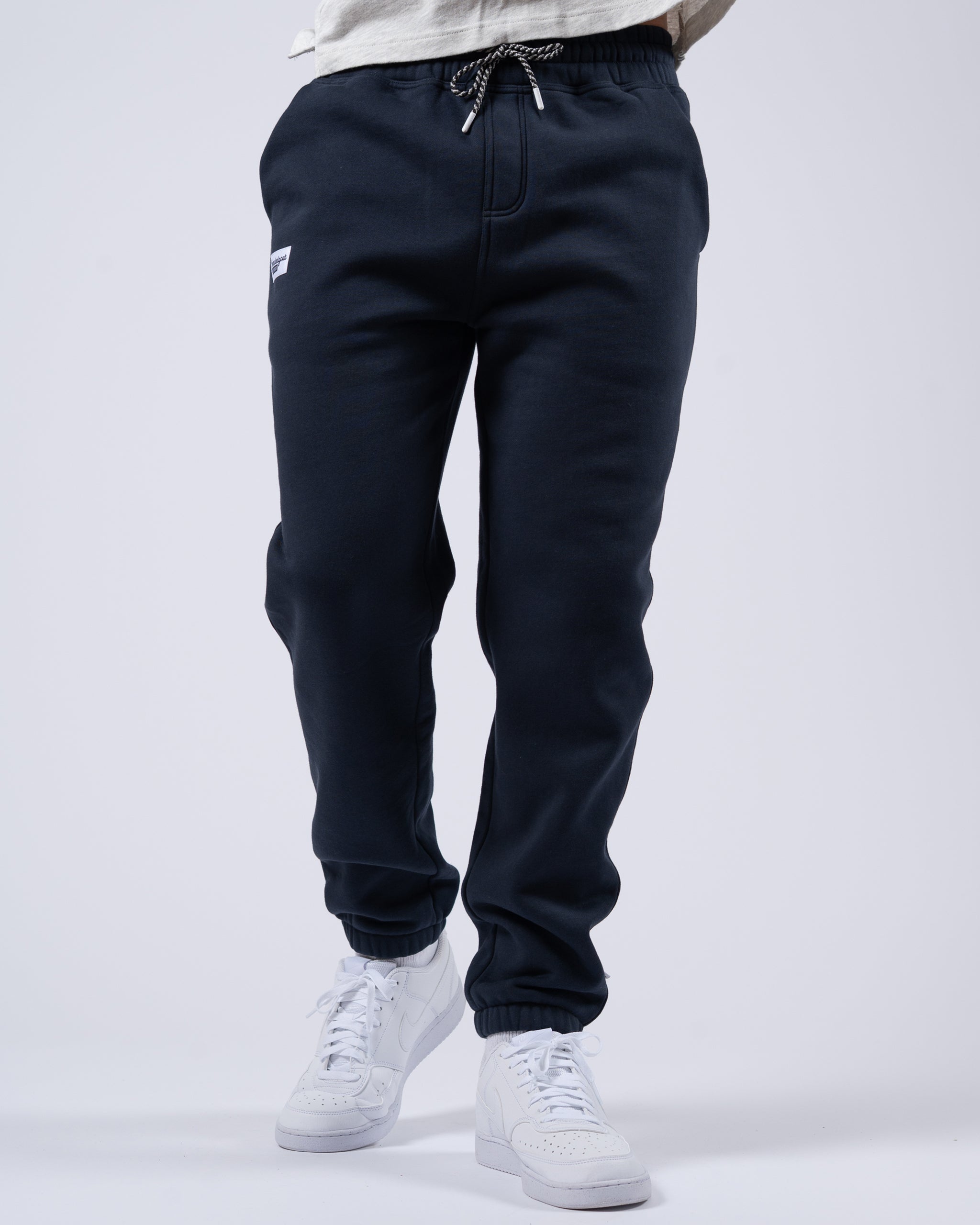 A person models the beardedgoat Men's Void Sweatpants in black, featuring a zippered back pocket, paired with white sneakers. Only the lower half is shown, set against a plain white background.
