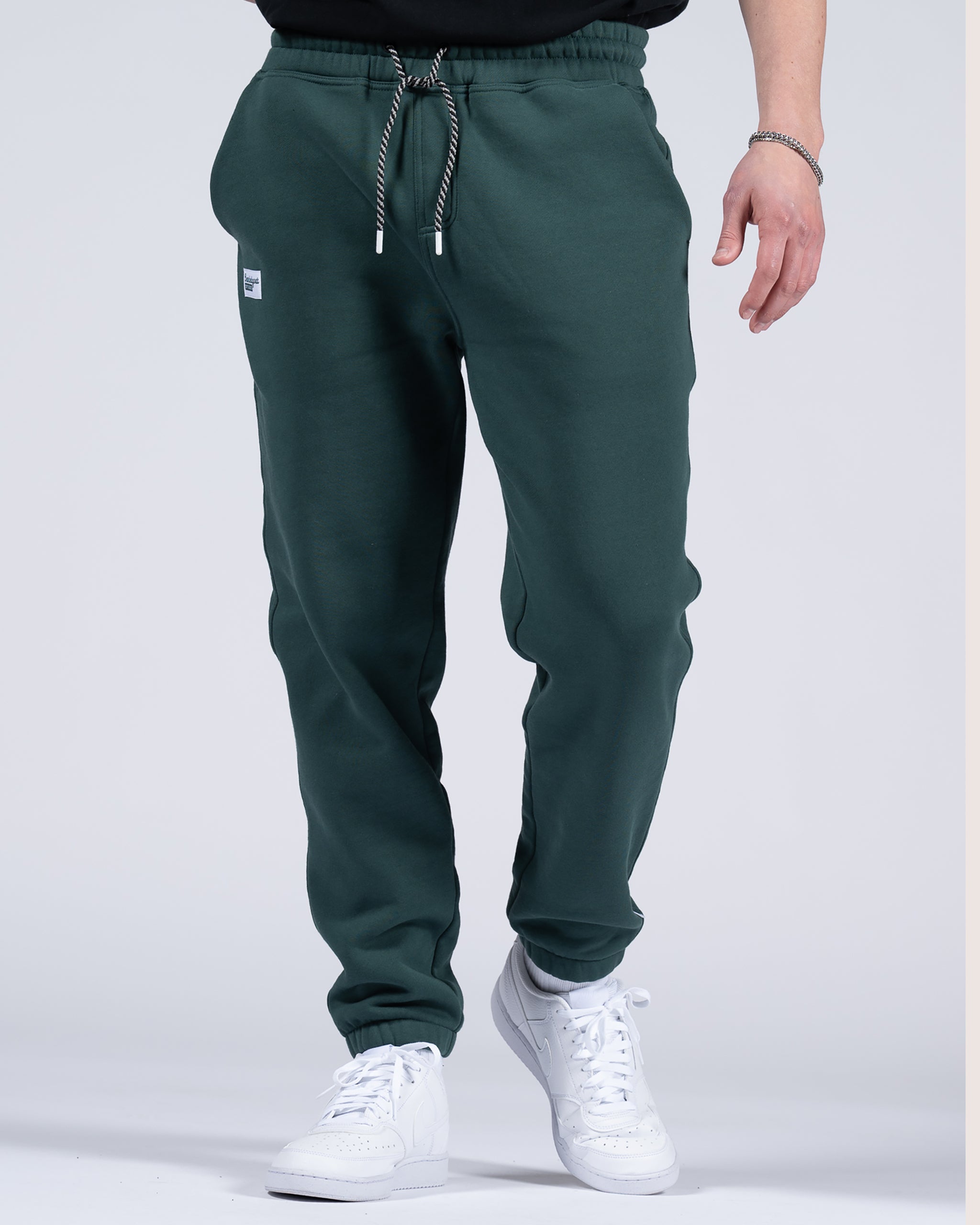 A person wearing beardedgoat Men's Void Sweatpants in dark green, featuring a drawstring waist and zipper back pocket, paired with white sneakers, is shown from the shoulders down against a light gray background.