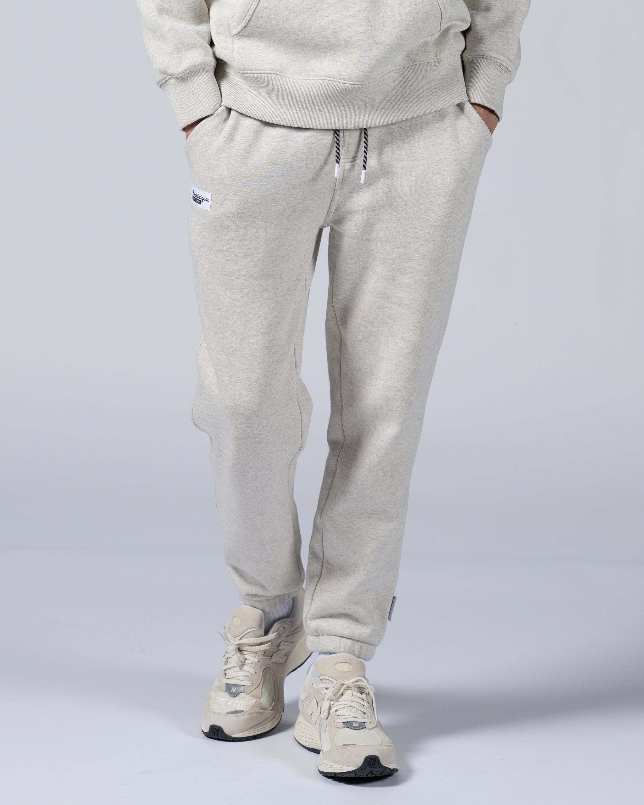 A person wears beardedgoat Men's Void Sweatpants in light gray with a drawstring, small upper leg patch, and zipper back pocket, hands in pockets against a plain background, paired with matching sneakers.