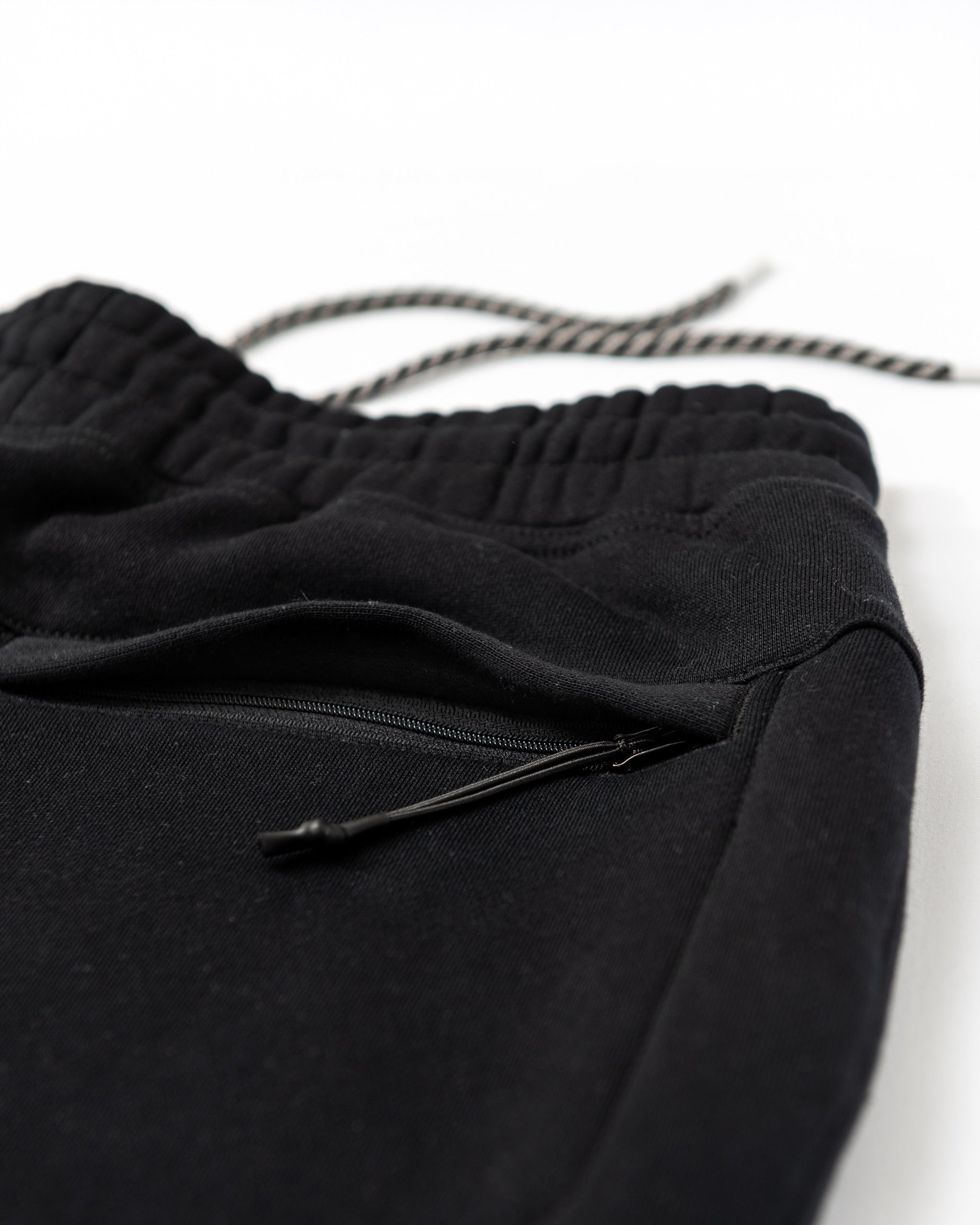 Close-up of beardedgoat Men's Void Sweatpants in black, featuring an elastic waistband, black-and-white drawstring, zippered side and back pockets—durable joggers shown against a white background.