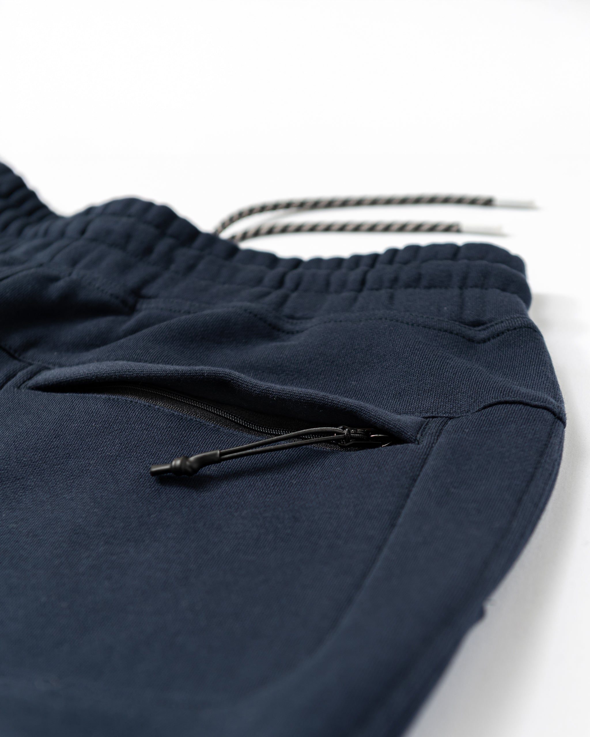Close-up of beardedgoat Men's Void Sweatpants in navy blue, featuring an elastic waistband, black and white drawstrings, zippered side and back pockets, and durable, soft, slightly textured fabric.