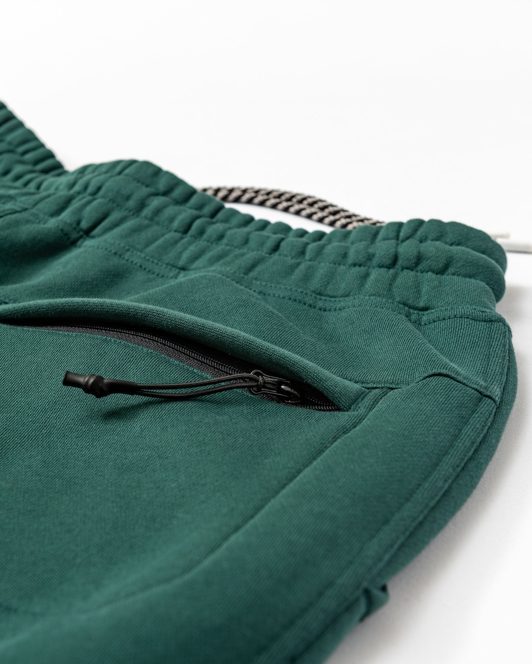 Close-up of green beardedgoat Men's Void Sweatpants, highlighting an elastic waistband with black-and-white drawstring, zippered side pocket, and soft, durable, slightly textured fabric.