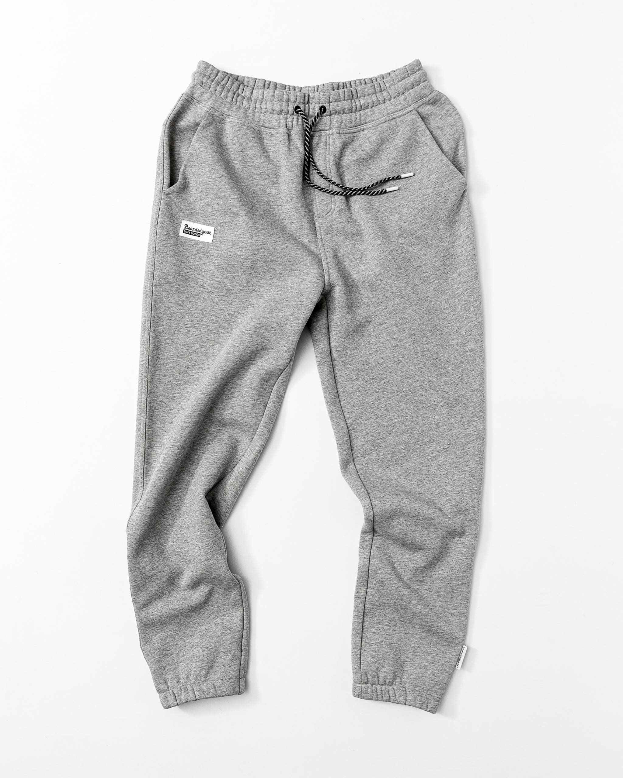 A pair of Men's Void Sweatpants by beardedgoat in light grey, featuring an elastic waistband, black-and-white drawstring, side and zipper back pockets, and a white patch label on the left thigh, laid flat on a white background.