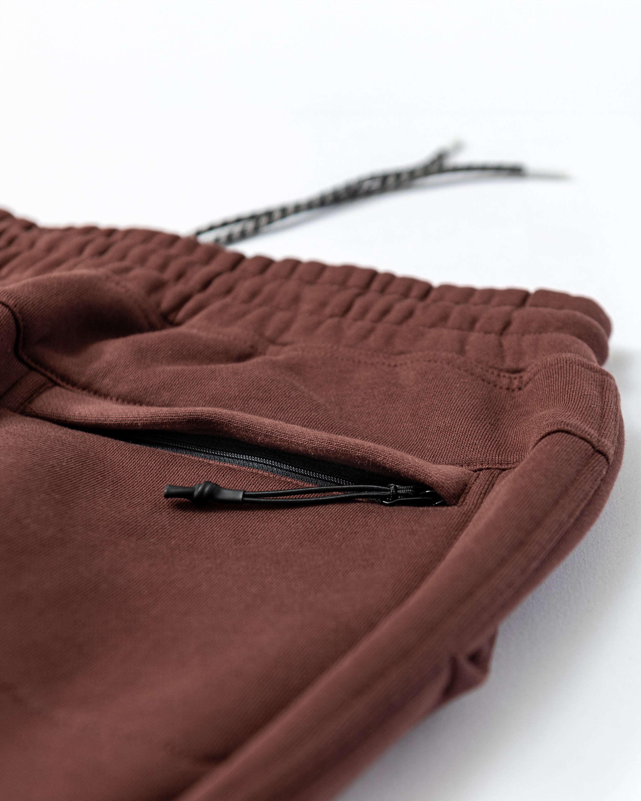 Close-up of beardedgoat Men's Void Sweatpants in maroon, featuring an elastic waistband with drawstring, zippered side and back pockets, displayed on a white surface. Durable and stylish for comfort.