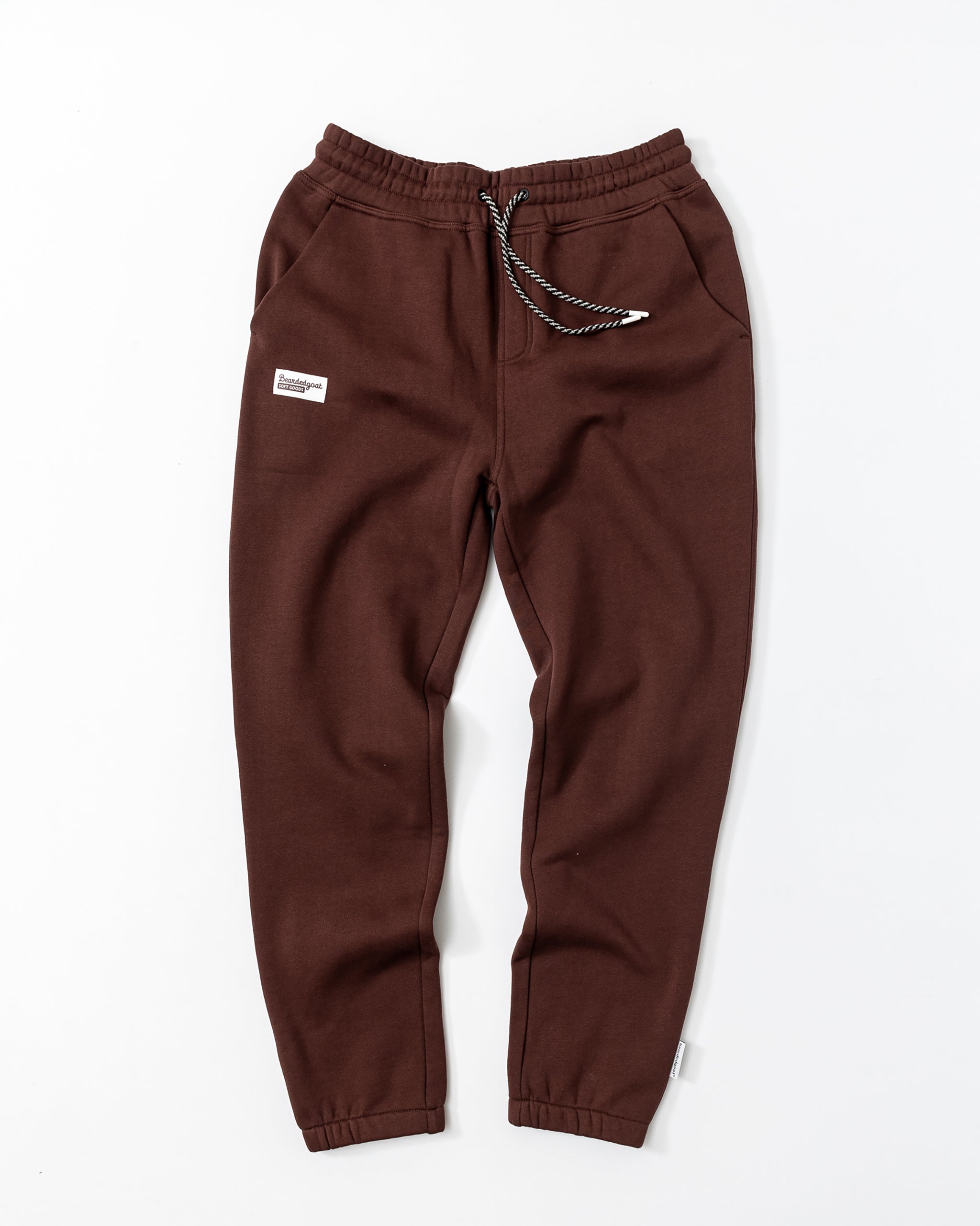 Men's Void Sweatpants by beardedgoat in brown, featuring an elastic waistband, black-and-white drawstring, side pockets, zipper back pocket, and a white rectangular patch on the left thigh, shown flat on a white background.