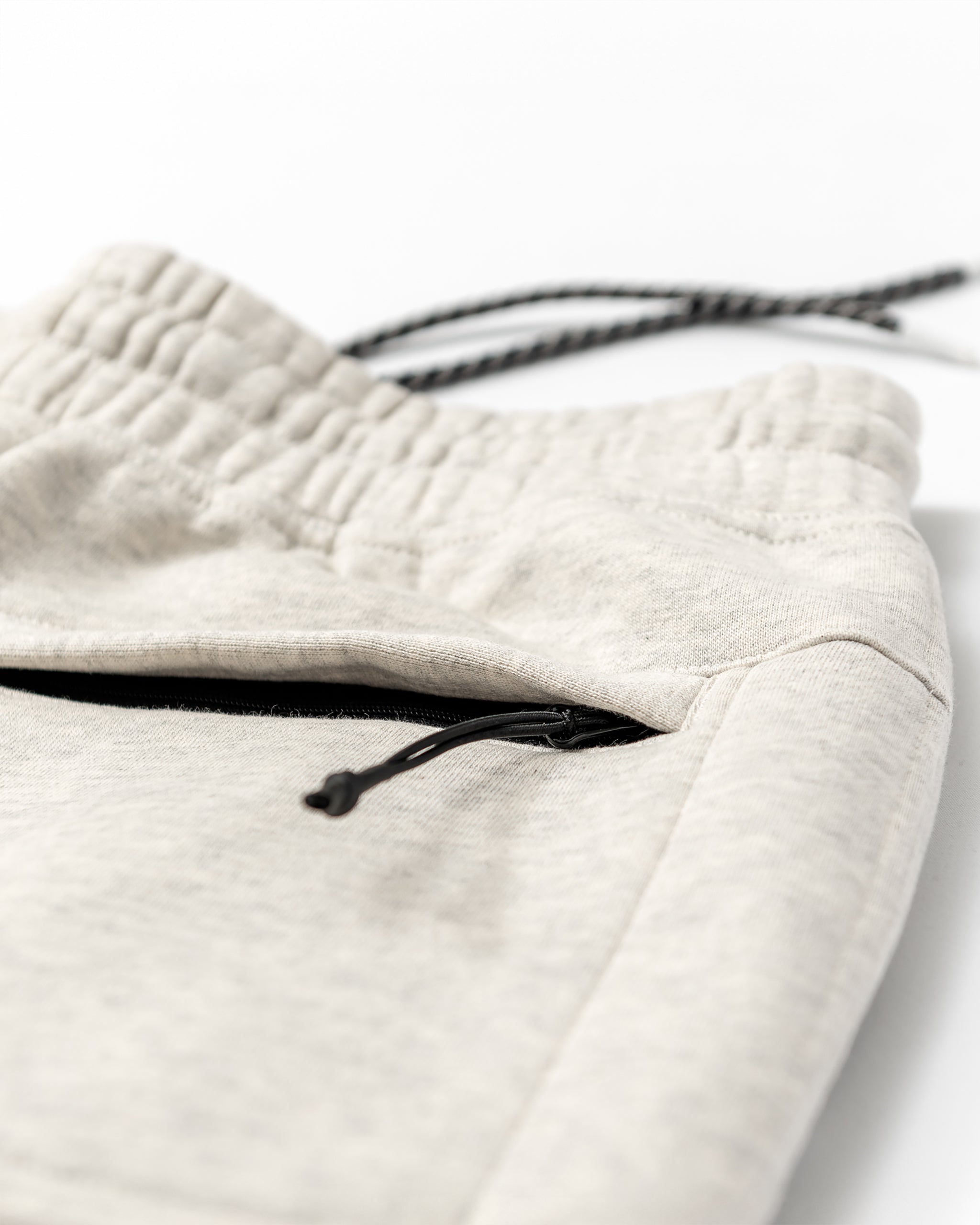 Close-up of beardedgoat Men's Void Sweatpants in light gray, featuring an elastic waistband, black drawstring, and a back zipper pocket with a black zipper pull, laid flat on a white surface.