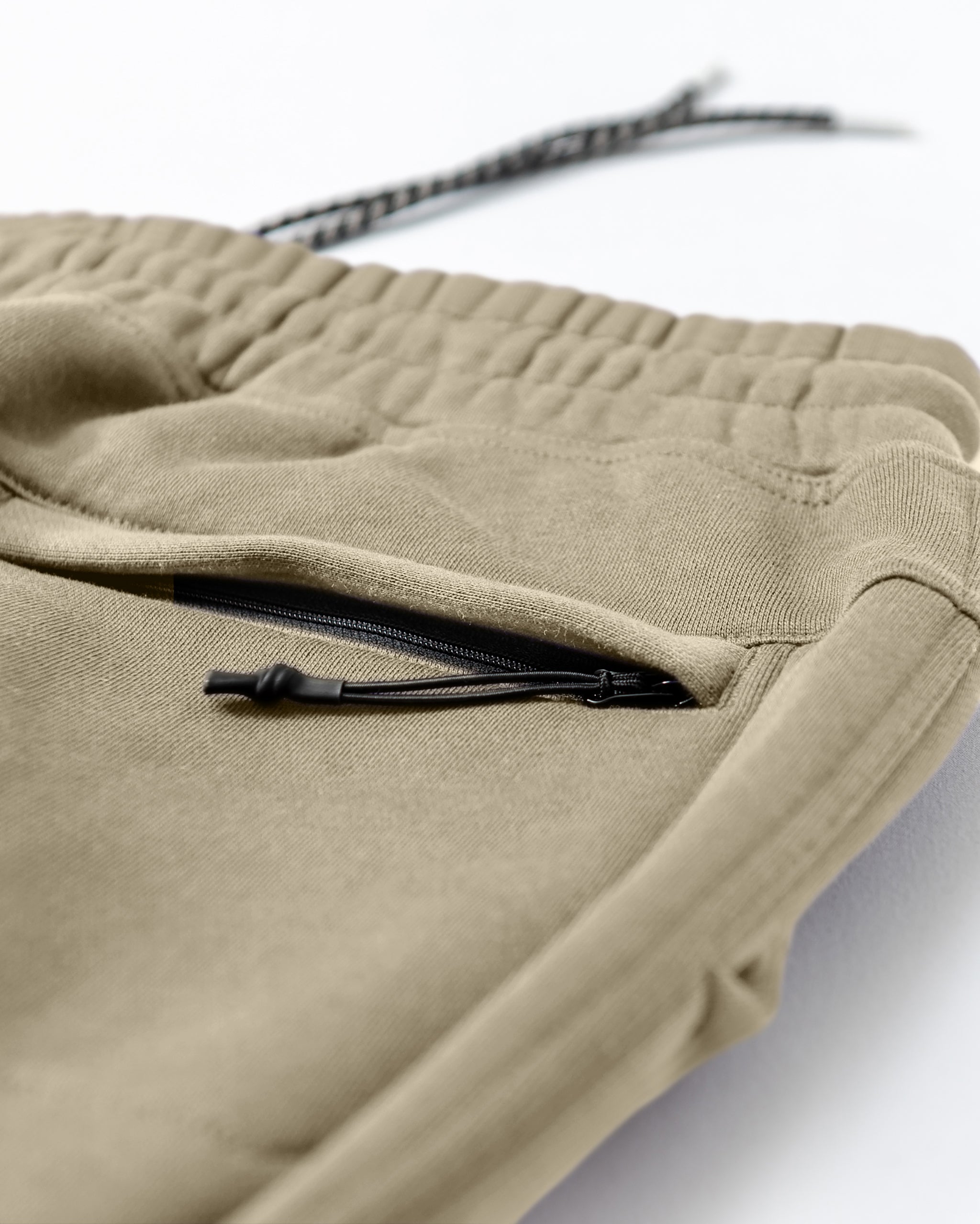 Close-up of tan beardedgoat Men's Void Sweatpants with an elastic waistband, a side pocket with black zipper, and a visible drawstring on a white surface—crafted from durable sweatpants material.