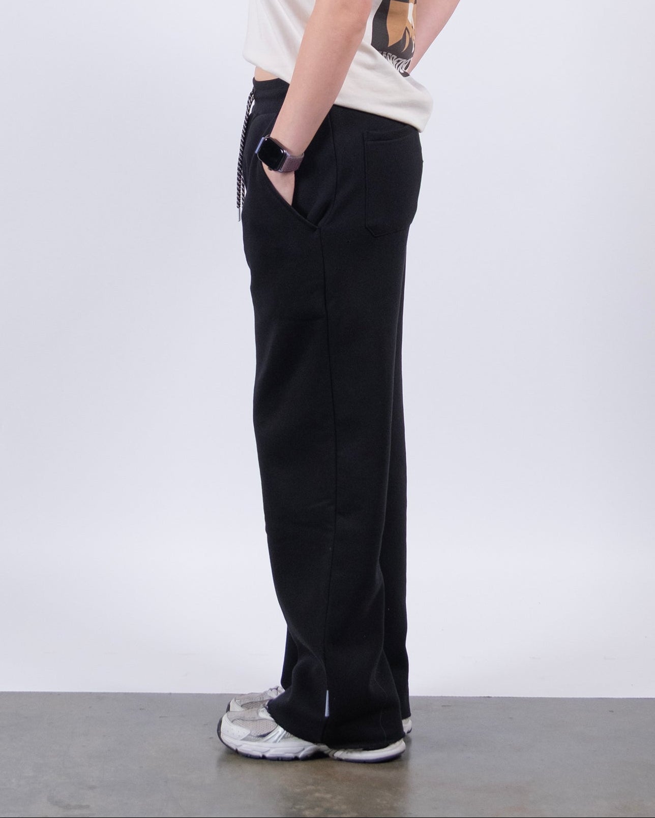A woman with long dark hair stands in profile against a white background, wearing a light t-shirt, beardedgoat Women’s Overnight Sweatpant made from heavyweight cotton fleece, white sneakers, and a smartwatch for a comfy loungewear look.