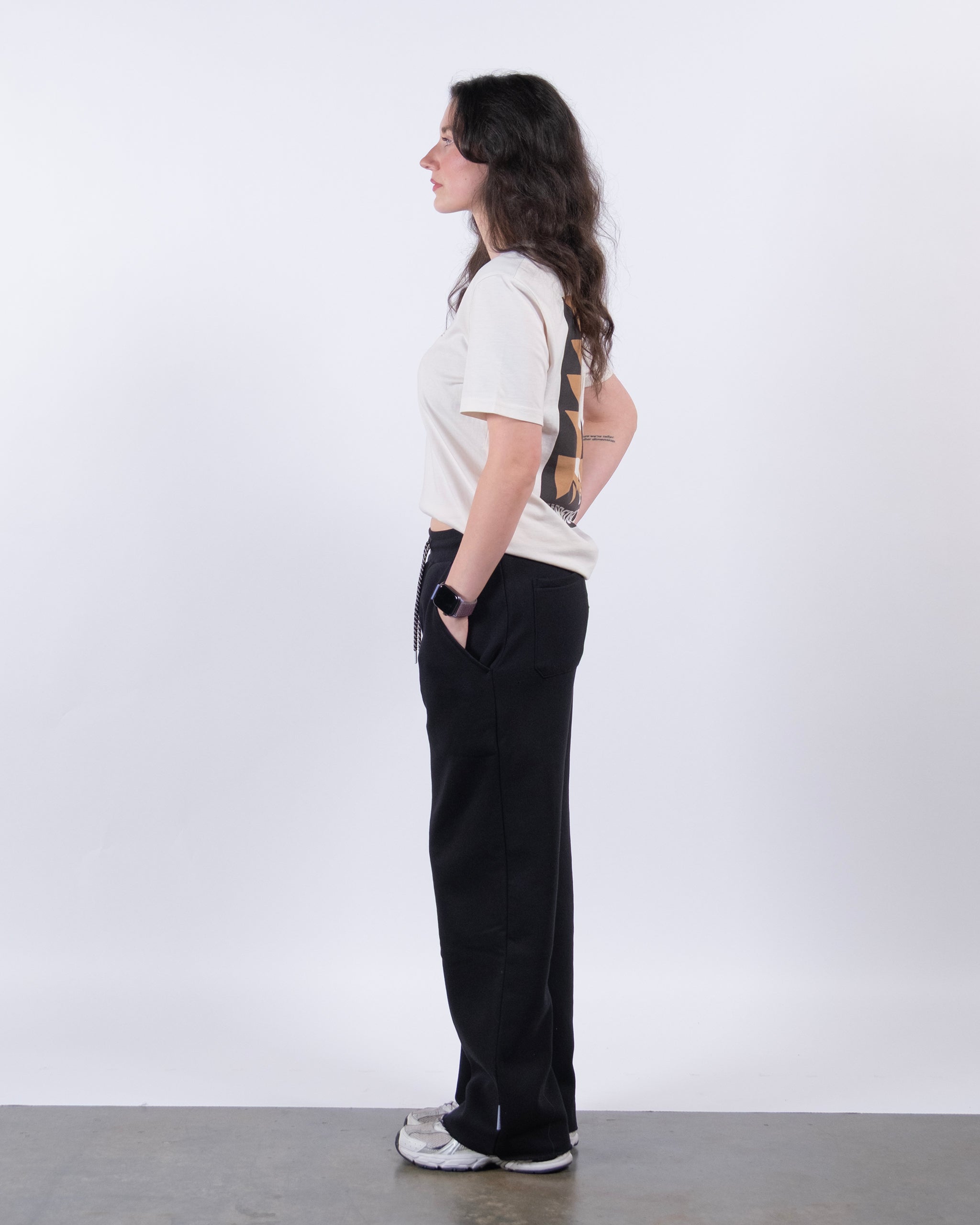 A woman with long dark hair stands in profile against a white background, wearing a light t-shirt, beardedgoat Women’s Overnight Sweatpant made from heavyweight cotton fleece, white sneakers, and a smartwatch for a comfy loungewear look.