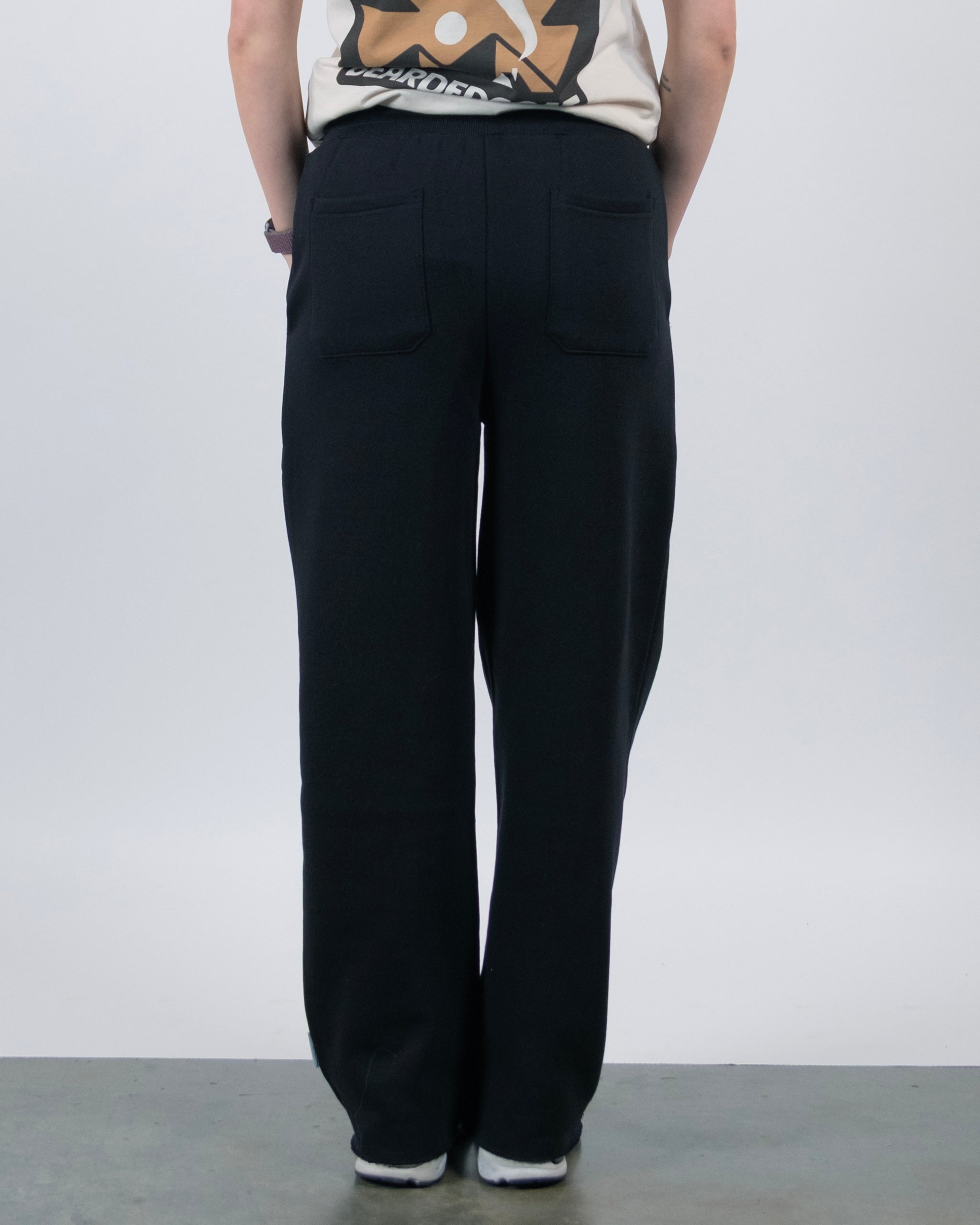 A person stands with their back to the camera, wearing a beige graphic tee and beardedgoat Women’s Overnight Sweatpant with back pockets. The relaxed outfit is on a gray floor against a plain white background.