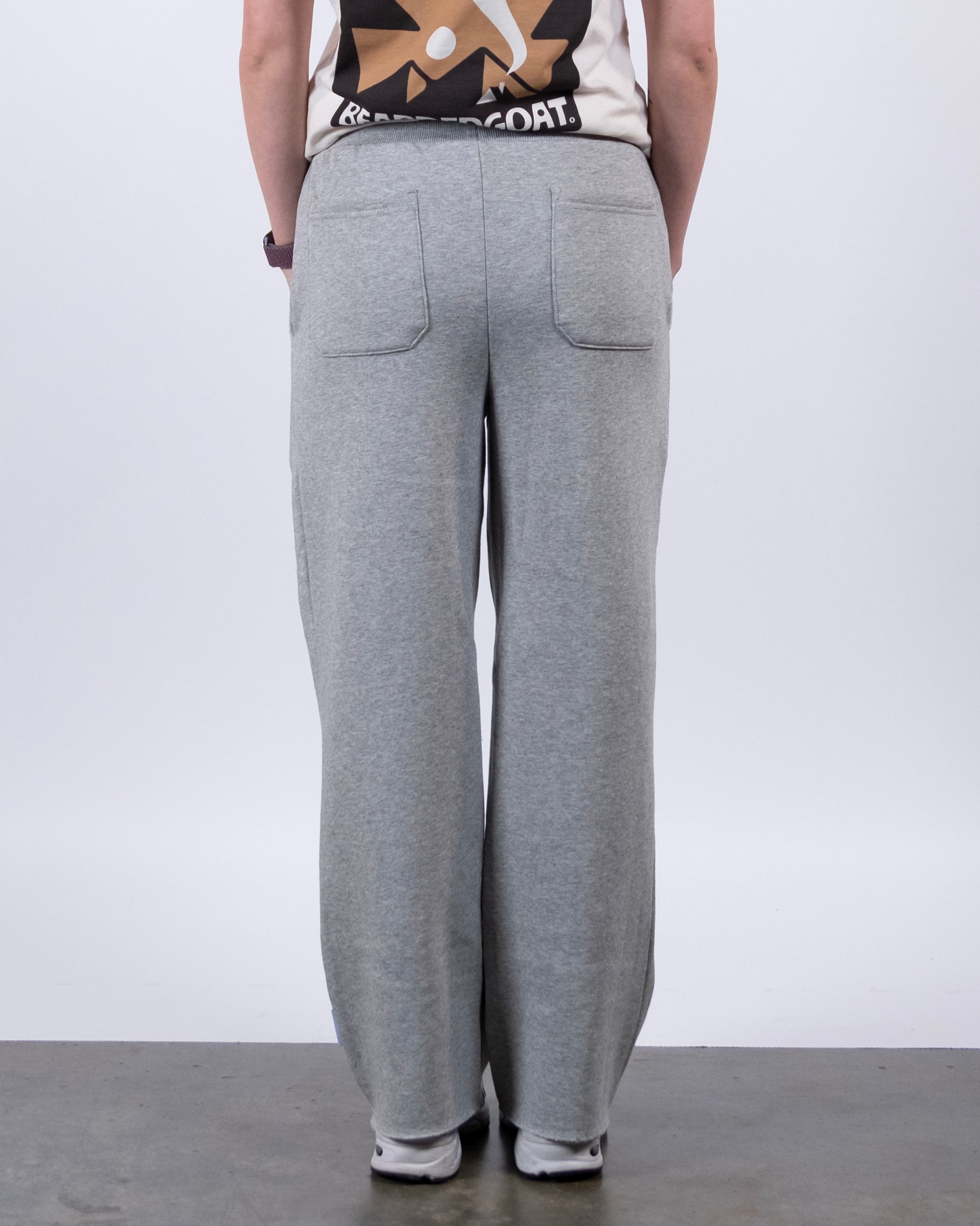 A person faces away from the camera, wearing beardedgoat’s Women’s Overnight Sweatpant in light gray and a beige graphic tee. The background and floor are plain gray.