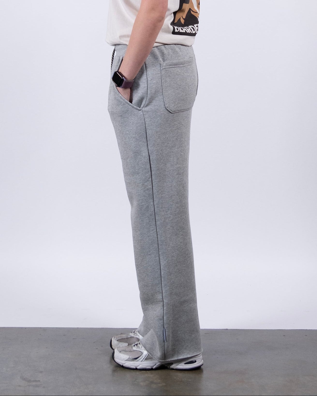 A woman with long dark hair stands in profile against a white background, wearing a light T-shirt, gray beardedgoat Women’s Overnight Sweatpant, and sneakers, with hands tucked casually into her pockets.